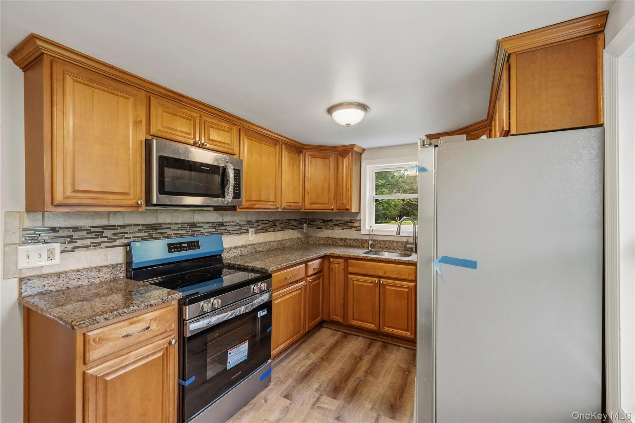 Kitchen featuring wood-finish cabinetry, granite countertops, tile backsplash, stainless steel microwave, and wood-finish flooring Kitchen featuring wood-finish cabinetry, granite countertops, tile backsplash, stainless steel microwave, and wood-finish flooring