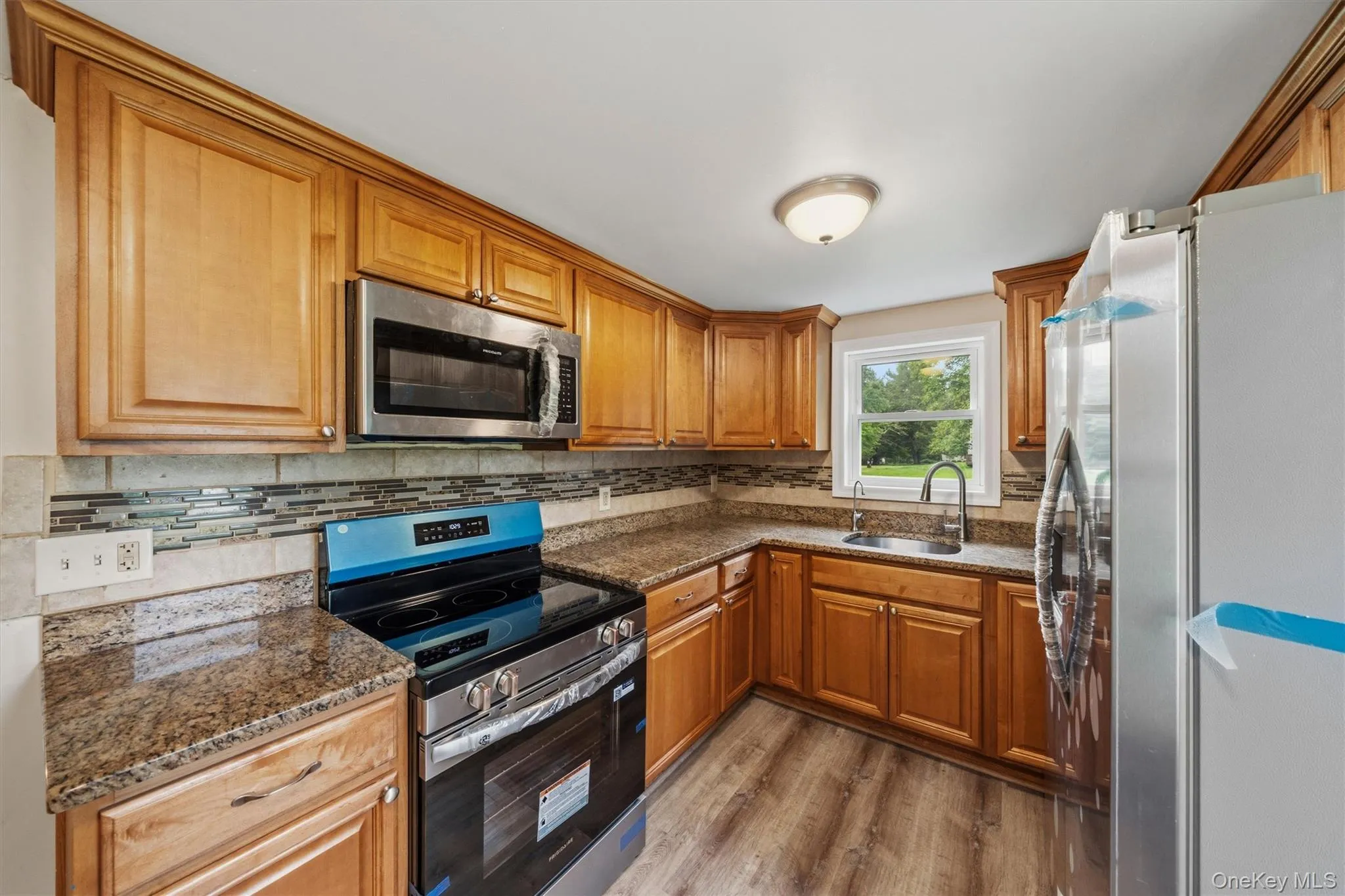 Kitchen featuring wood-finish cabinetry, granite-style countertops, stainless steel appliances, a multi-tone tile backsplash, and wood-look flooring Kitchen featuring wood-finish cabinetry, granite-style countertops, stainless steel appliances, a multi-tone tile backsplash, and wood-look flooring