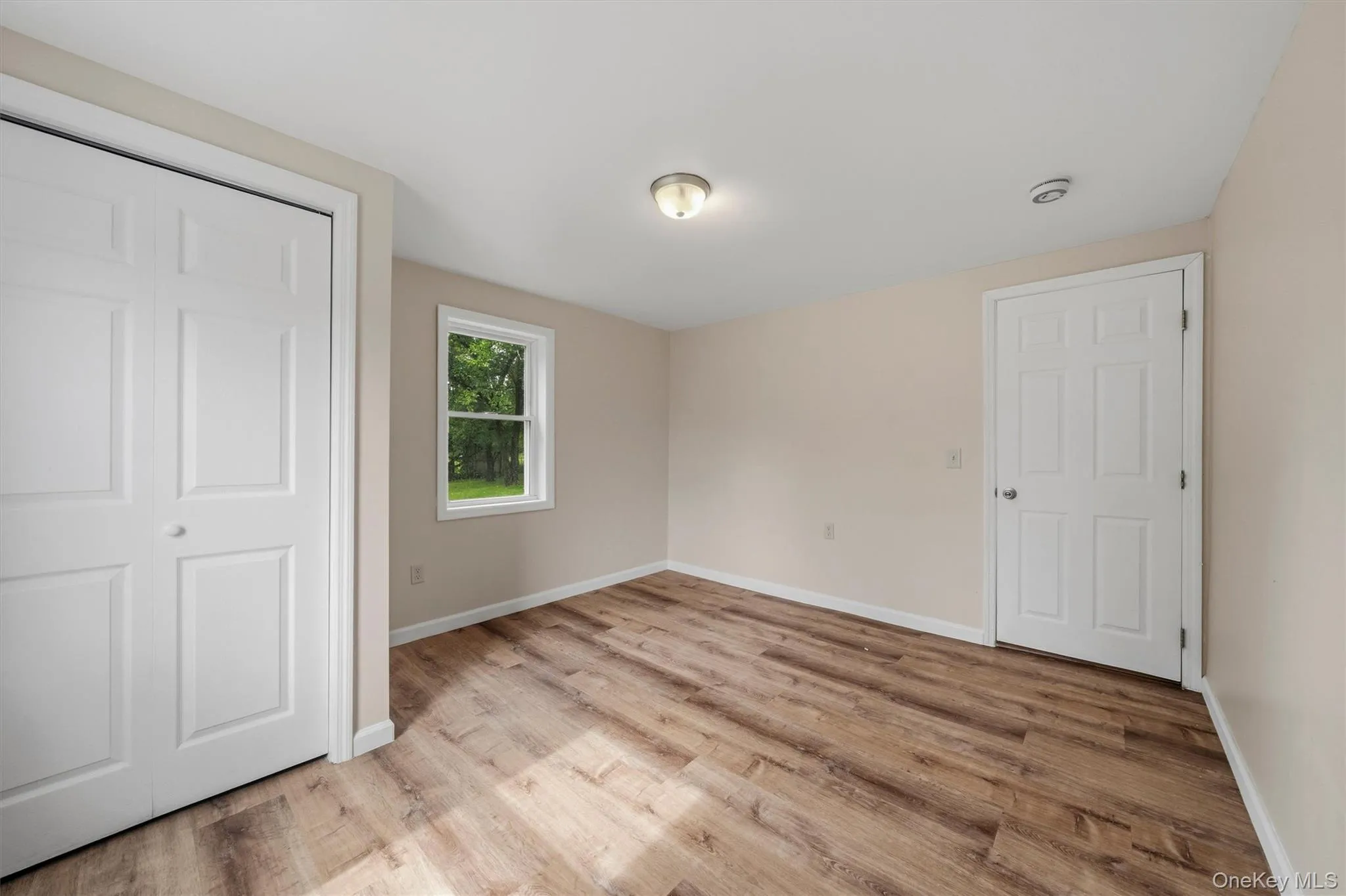 Wood-finish flooring, white bi-fold closet doors, white window trim, and a ceiling-mounted light fixture Wood-finish flooring, white bi-fold closet doors, white window trim, and a ceiling-mounted light fixture