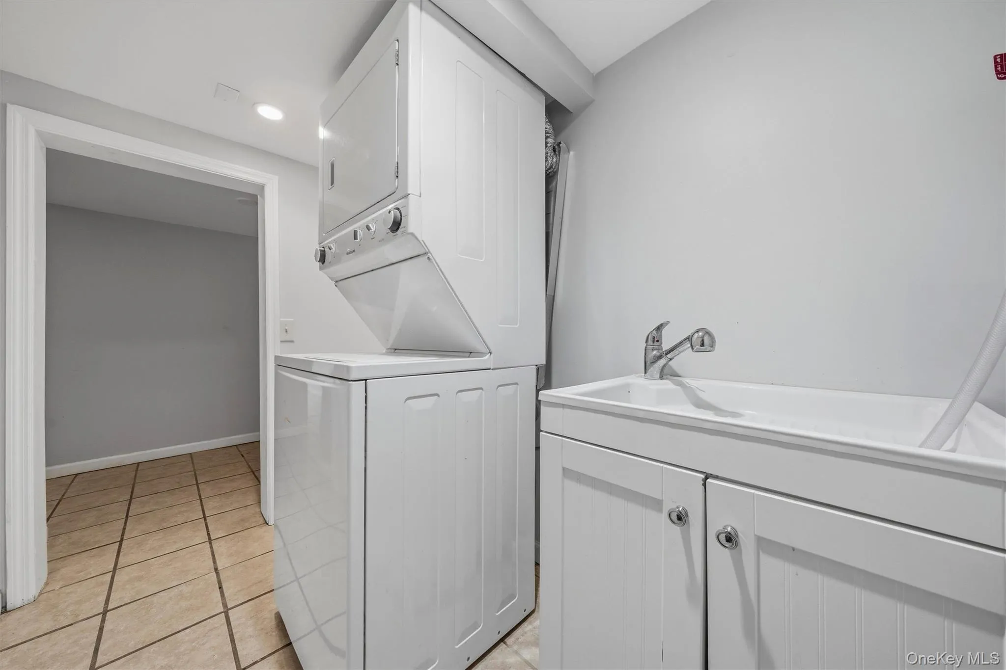 Laundry area featuring a stackable washer and dryer, utility sink, white base cabinetry, and tile flooring Laundry area featuring a stackable washer and dryer, utility sink, white base cabinetry, and tile flooring
