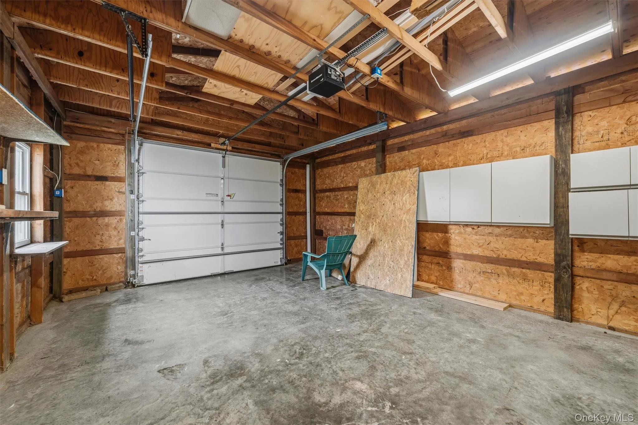 Concrete flooring with an overhead garage door, exposed wood beam ceiling, built-in wall cabinetry, and a single window Concrete flooring with an overhead garage door, exposed wood beam ceiling, built-in wall cabinetry, and a single window