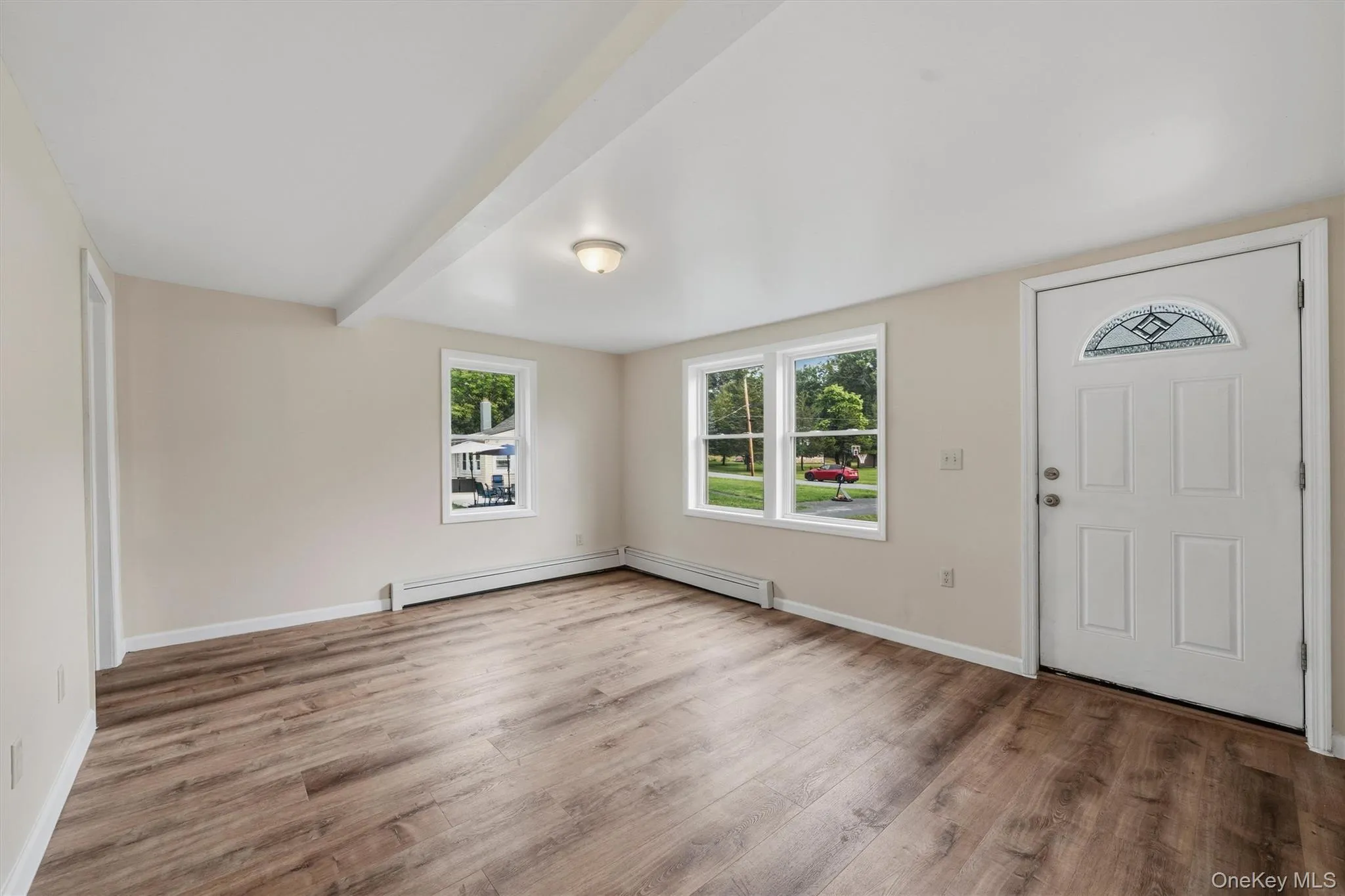 Spacious room featuring wood-finish flooring, neutral-toned walls, multiple windows, and a front entry door with an arched decorative glass insert Spacious room featuring wood-finish flooring, neutral-toned walls, multiple windows, and a front entry door with an arched decorative glass insert
