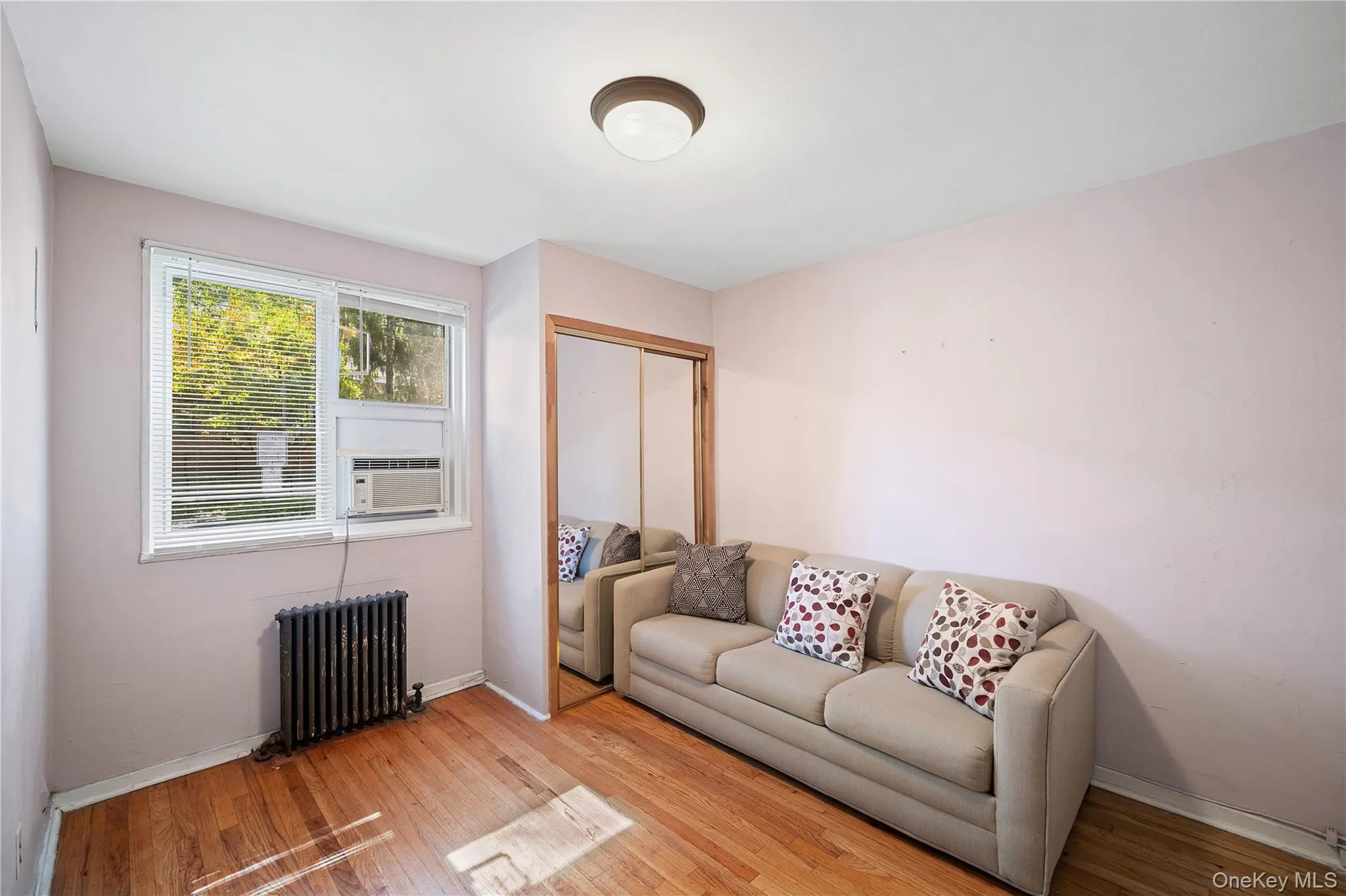 Wood-finish flooring with a window AC unit, ceiling-mounted light fixture, and mirrored closet doors Wood-finish flooring with a window AC unit, ceiling-mounted light fixture, and mirrored closet doors
