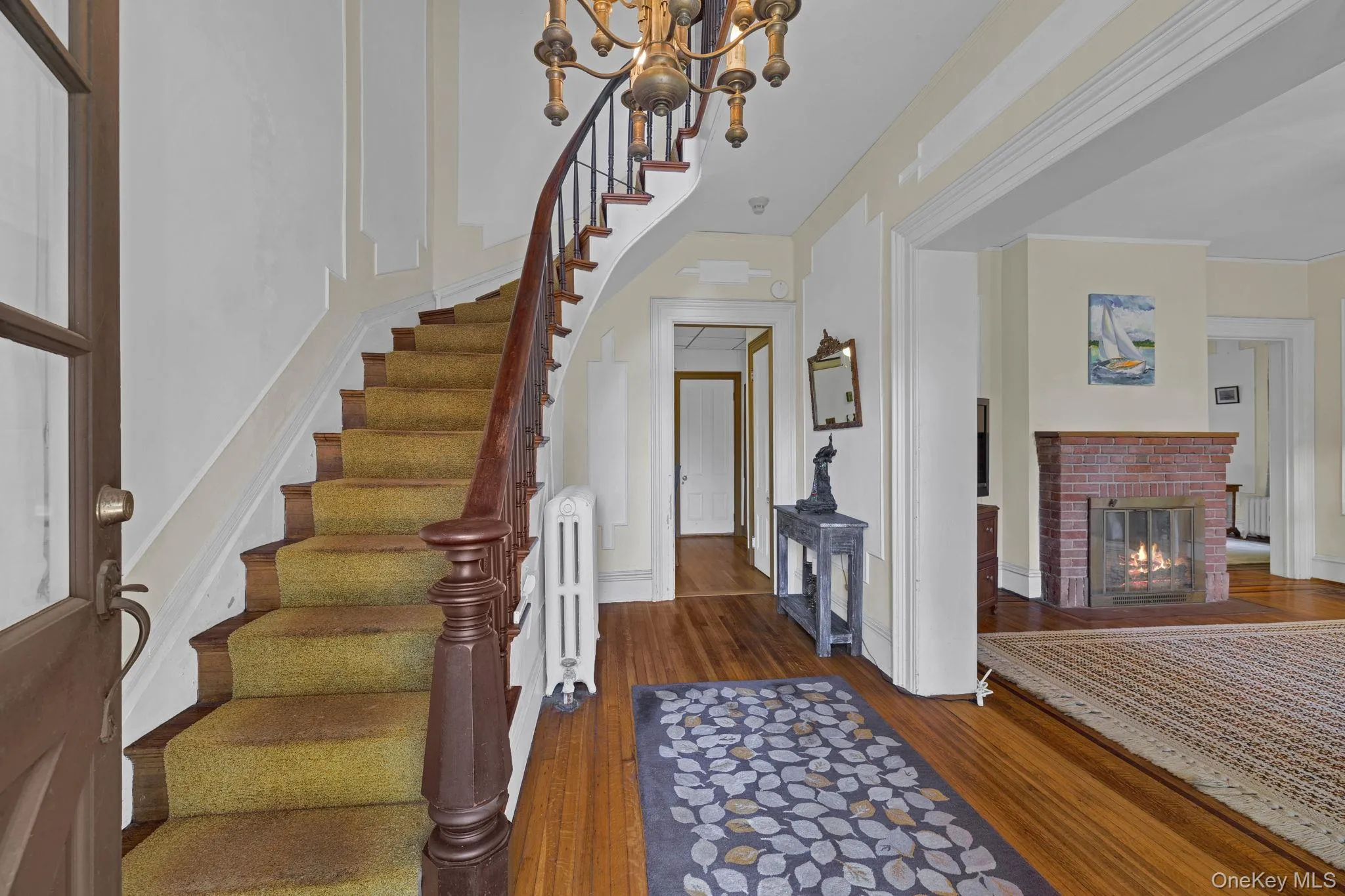 Foyer featuring dark wood-style floors, radiator, a brick fireplace, stairs, and a chandelier Foyer featuring dark wood-style floors, radiator, a brick fireplace, stairs, and a chandelier
