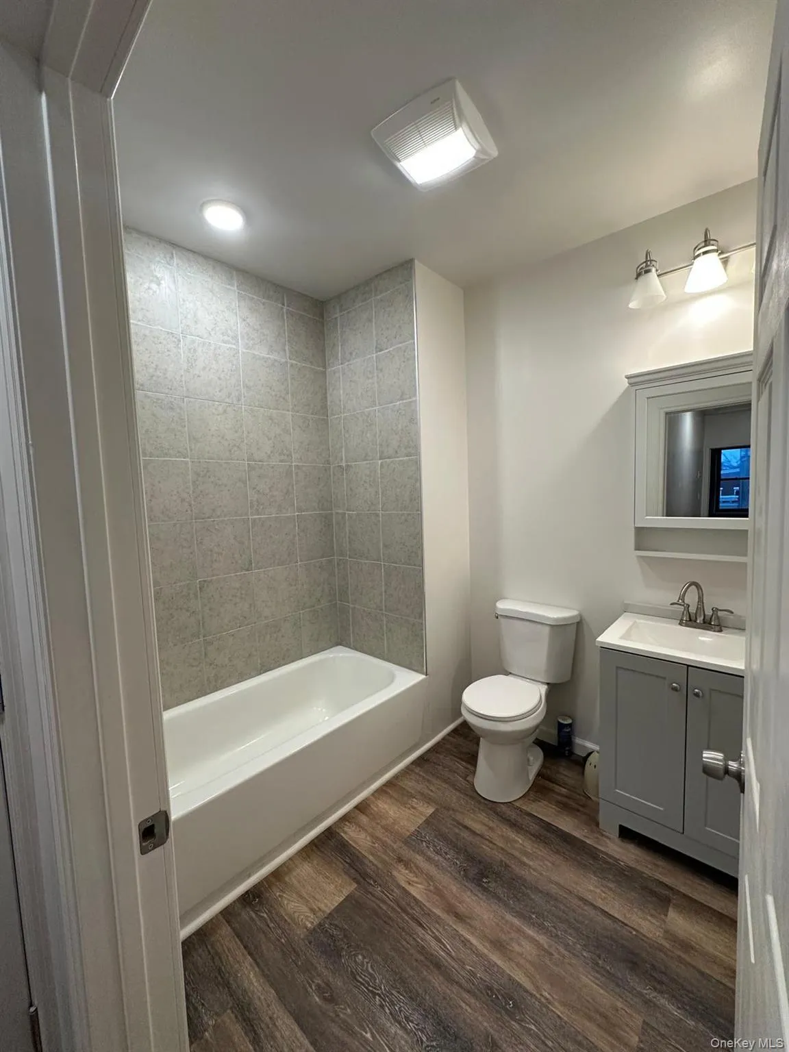Bathroom featuring a white bathtub, tiled shower surround, wood-finish flooring, a vanity with a white sink, and a mirrored medicine cabinet Bathroom featuring a white bathtub, tiled shower surround, wood-finish flooring, a vanity with a white sink, and a mirrored medicine cabinet