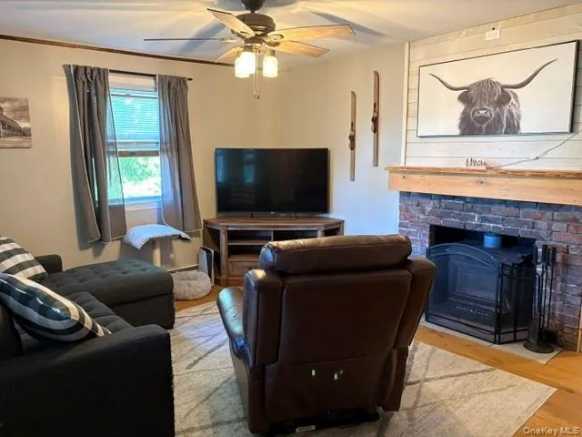 Brick fireplace with a wood mantel, wood-finish flooring, ceiling fan, and a window with blinds Brick fireplace with a wood mantel, wood-finish flooring, ceiling fan, and a window with blinds
