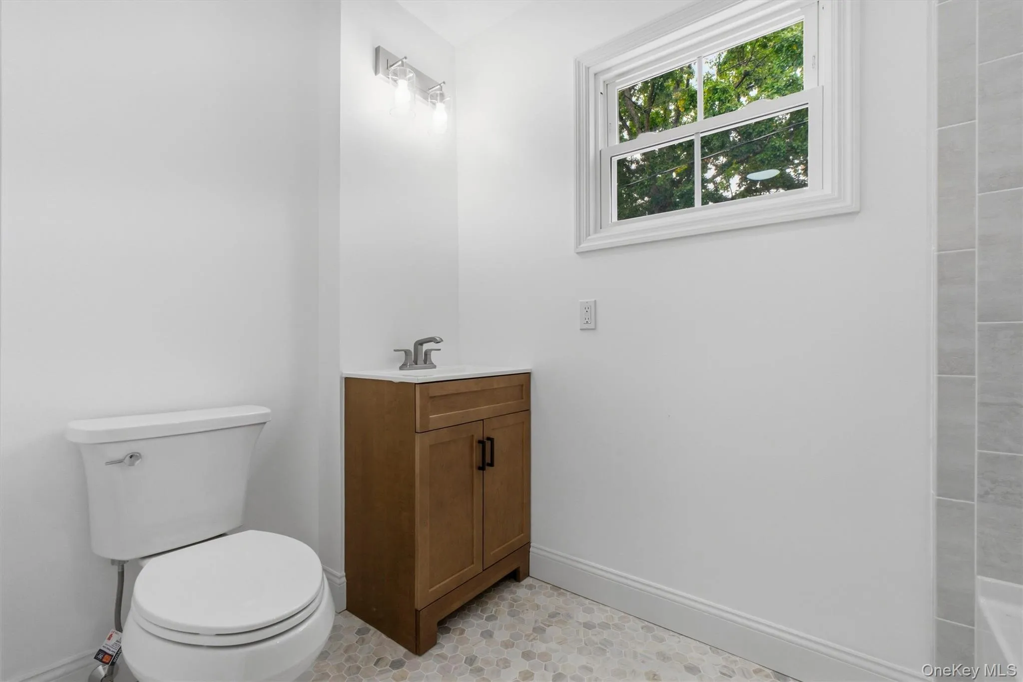 White wall paint throughout, light wood-finish vanity with a white countertop, window with white trim, and a mosaic tile floor White wall paint throughout, light wood-finish vanity with a white countertop, window with white trim, and a mosaic tile floor
