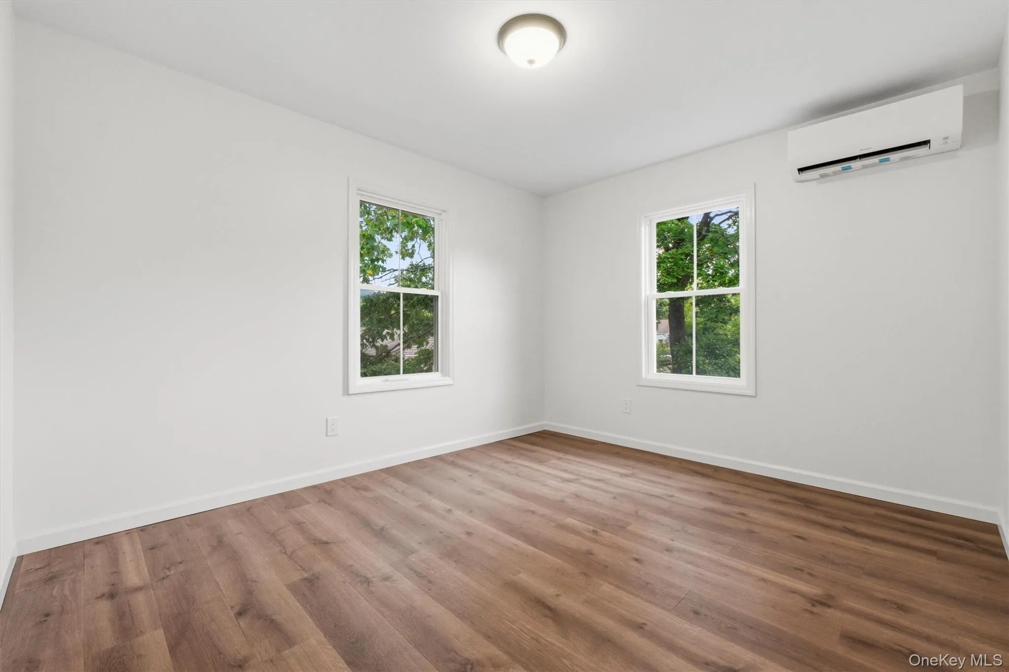 Wood-finish flooring, white walls, two windows, ceiling-mounted light fixture, and a wall-mounted air conditioning unit Wood-finish flooring, white walls, two windows, ceiling-mounted light fixture, and a wall-mounted air conditioning unit