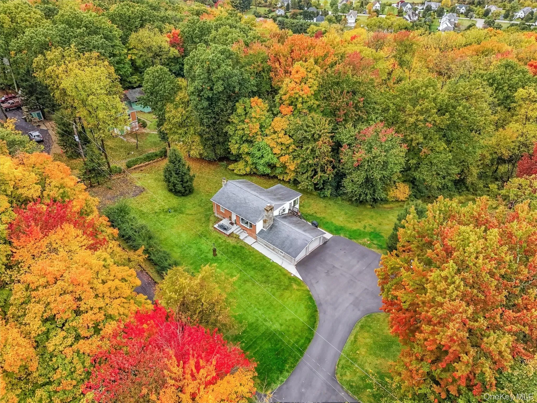Aerial view highlighting the home, attached two-car garage, expansive paved driveway, grassy yard, and mature trees with colorful fall foliage. Aerial view highlighting the home, attached two-car garage, expansive paved driveway, grassy yard, and mature trees with colorful fall foliage.