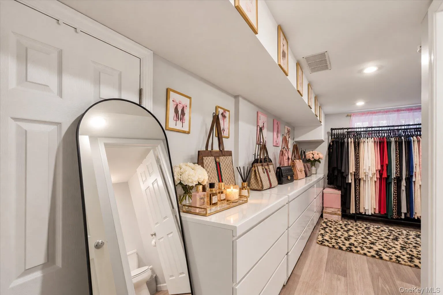 Virtually staged clothes and decluttered. Spacious walk-in closet featuring white drawer storage units, clothing racks, a full-length mirror, and wood-look flooring. Virtually staged clothes and decluttered. Spacious walk-in closet featuring white drawer storage units, clothing racks, a full-length mirror, and wood-look flooring.
