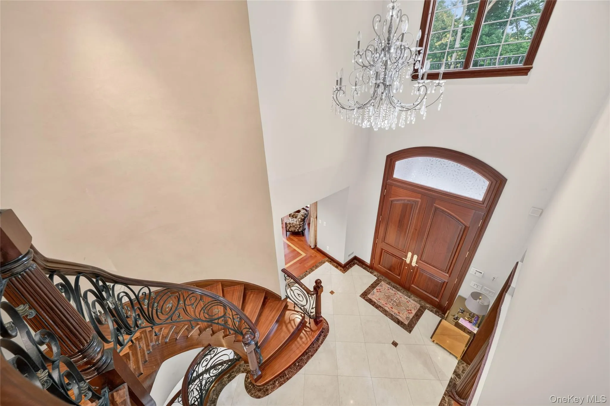 Grand foyer featuring a crystal chandelier, arched double entry doors, a large transom window, a curved staircase with an ornate wrought iron railing, and tile flooring with decorative inlay Grand foyer featuring a crystal chandelier, arched double entry doors, a large transom window, a curved staircase with an ornate wrought iron railing, and tile flooring with decorative inlay