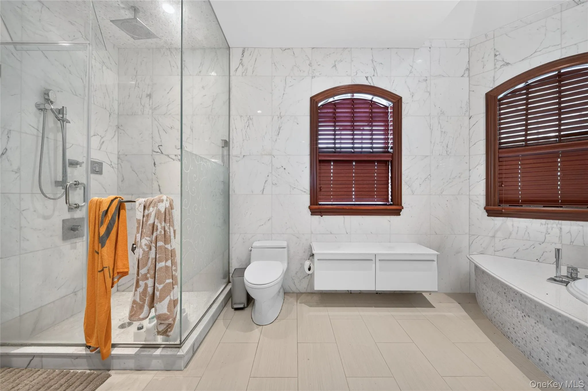 Glass-enclosed shower with multiple showerheads, white tile walls, two arched windows with wood-tone blinds, and a built-in bathtub with a white countertop Glass-enclosed shower with multiple showerheads, white tile walls, two arched windows with wood-tone blinds, and a built-in bathtub with a white countertop