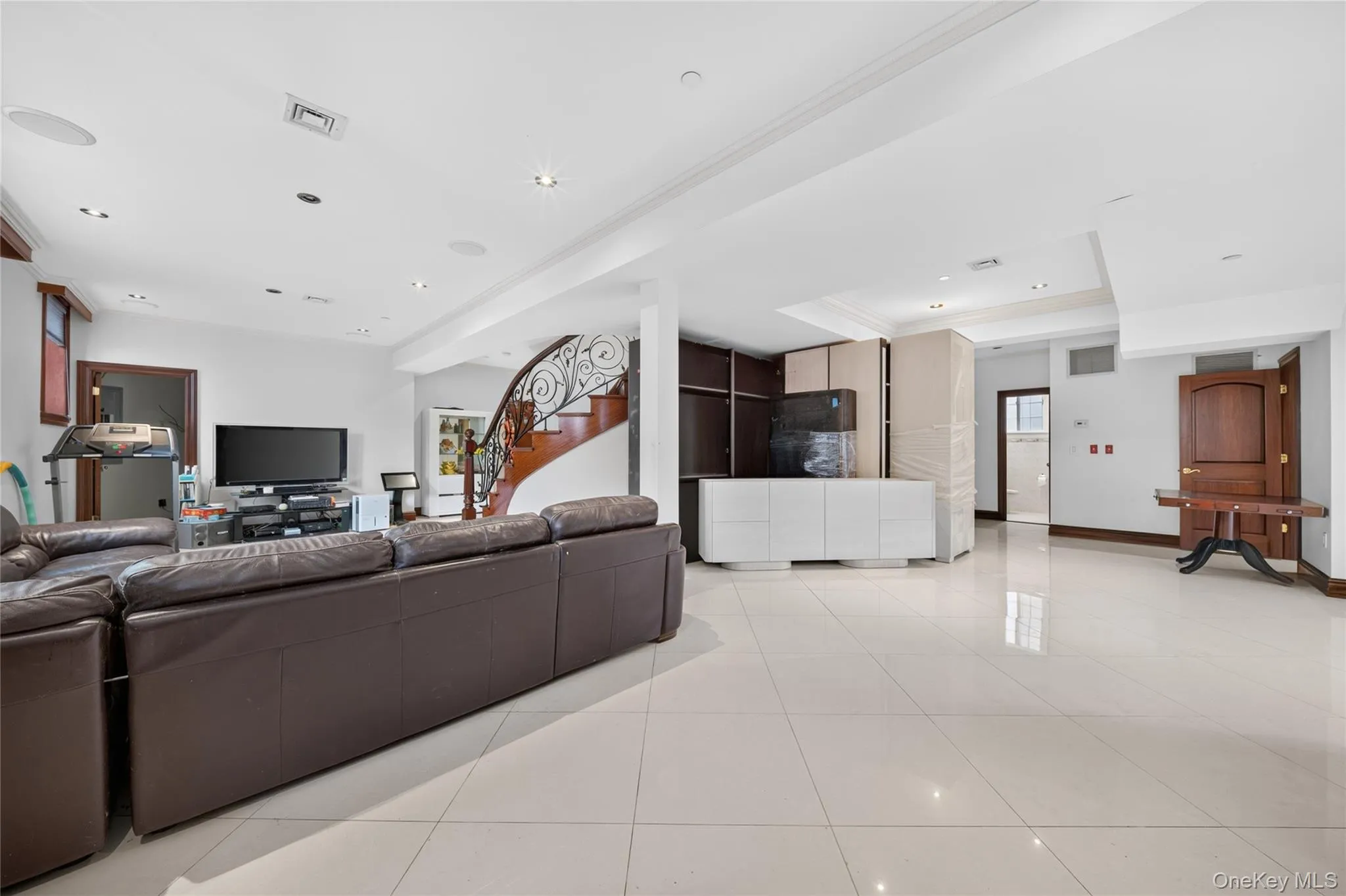 Spacious open-concept room featuring large format tile flooring, recessed lighting, a built-in media cabinet, and a curved staircase with an ornate wrought iron railing Spacious open-concept room featuring large format tile flooring, recessed lighting, a built-in media cabinet, and a curved staircase with an ornate wrought iron railing