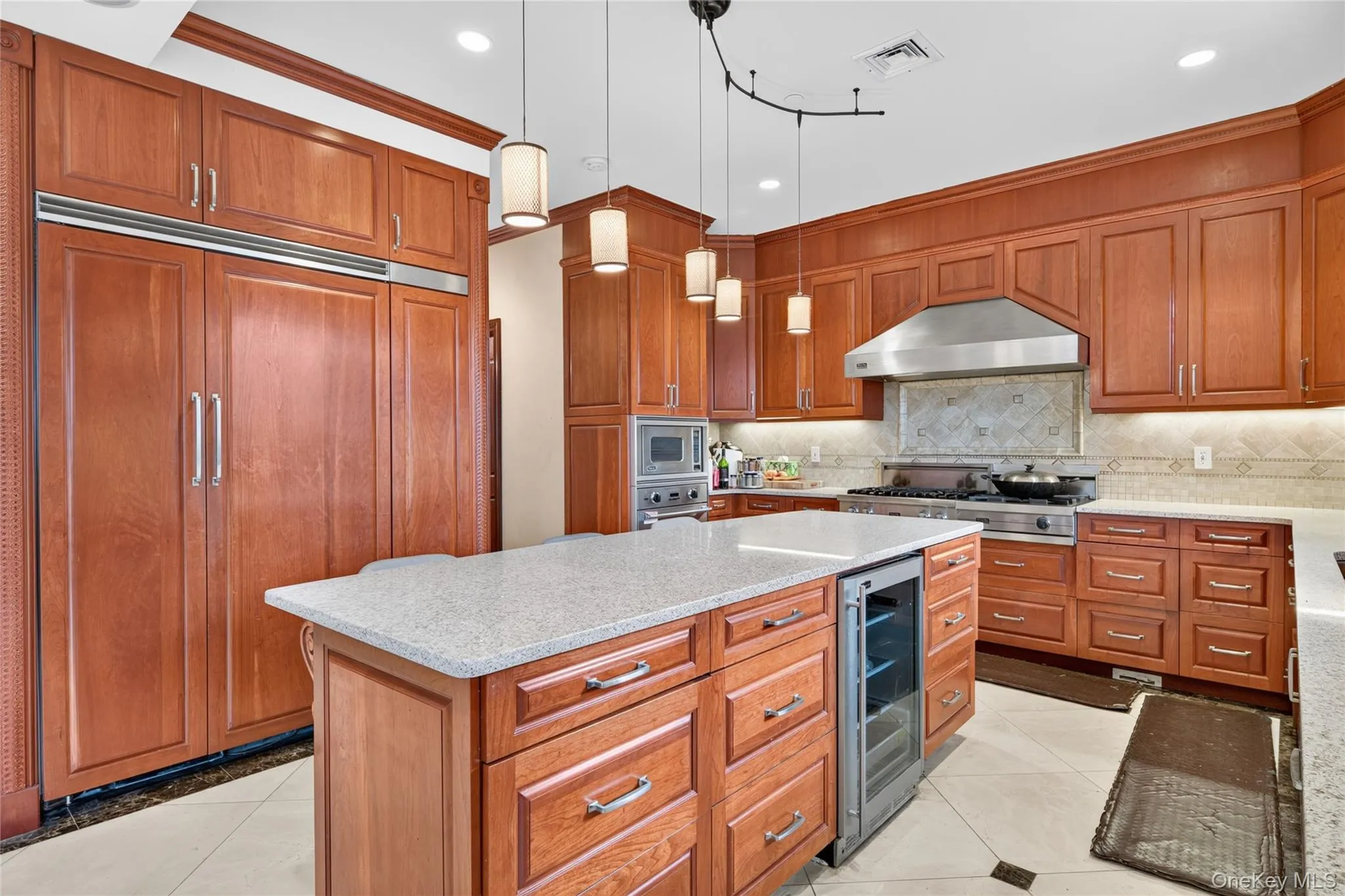 Custom wood cabinetry, stainless steel appliances, central island with integrated beverage refrigerator, light-toned countertops, and recessed and pendant lighting Custom wood cabinetry, stainless steel appliances, central island with integrated beverage refrigerator, light-toned countertops, and recessed and pendant lighting