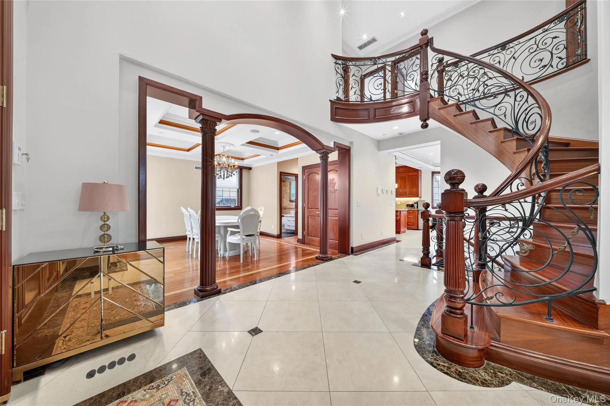Grand entryway featuring a curved staircase with ornate ironwork and wood-finish treads, a coffered ceiling, and a formal dining space with hardwood flooring Grand entryway featuring a curved staircase with ornate ironwork and wood-finish treads, a coffered ceiling, and a formal dining space with hardwood flooring