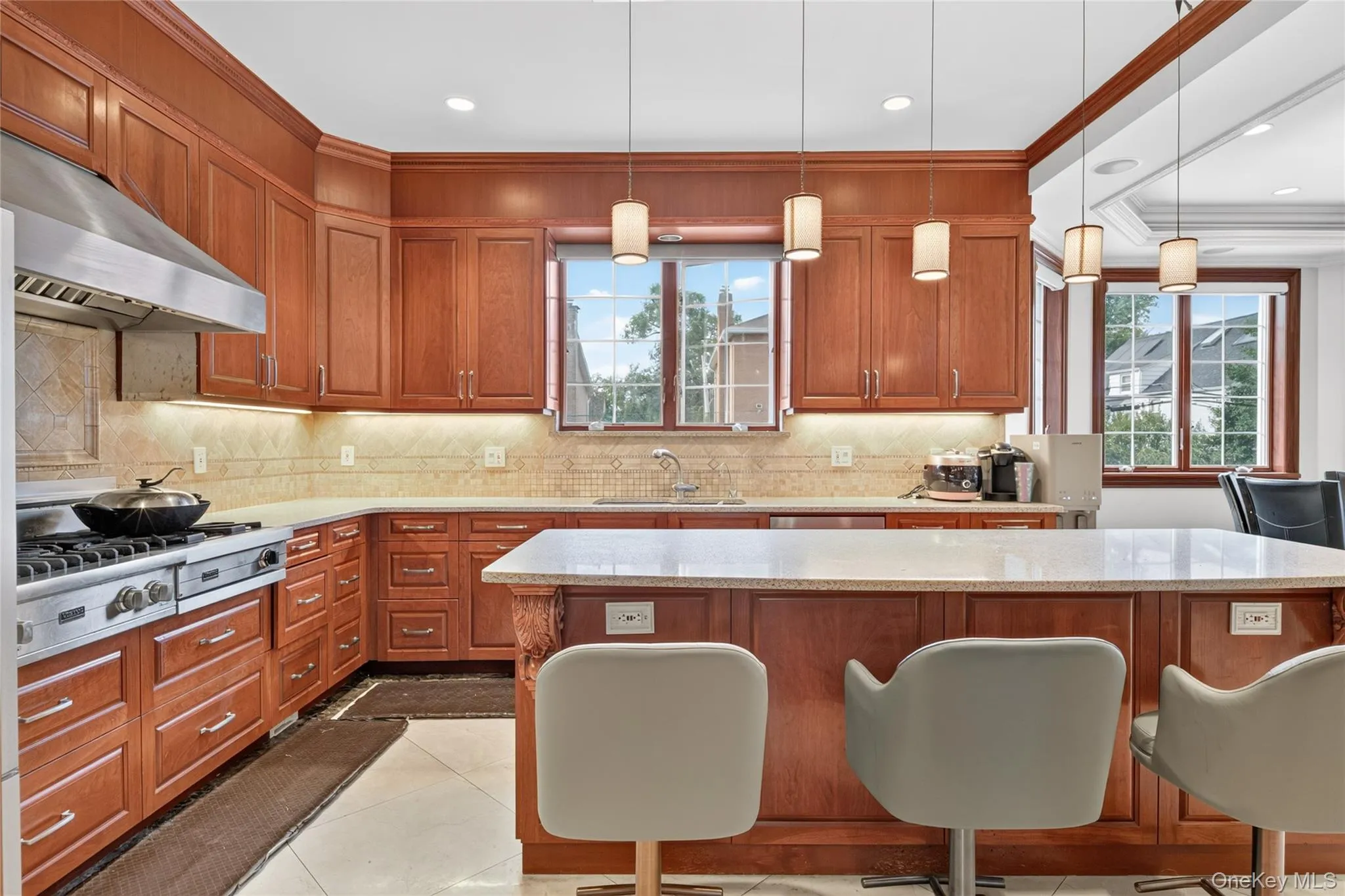 Wood cabinetry, light-toned backsplash, stainless steel range with hood, island with light-toned countertop, and pendant lighting Wood cabinetry, light-toned backsplash, stainless steel range with hood, island with light-toned countertop, and pendant lighting