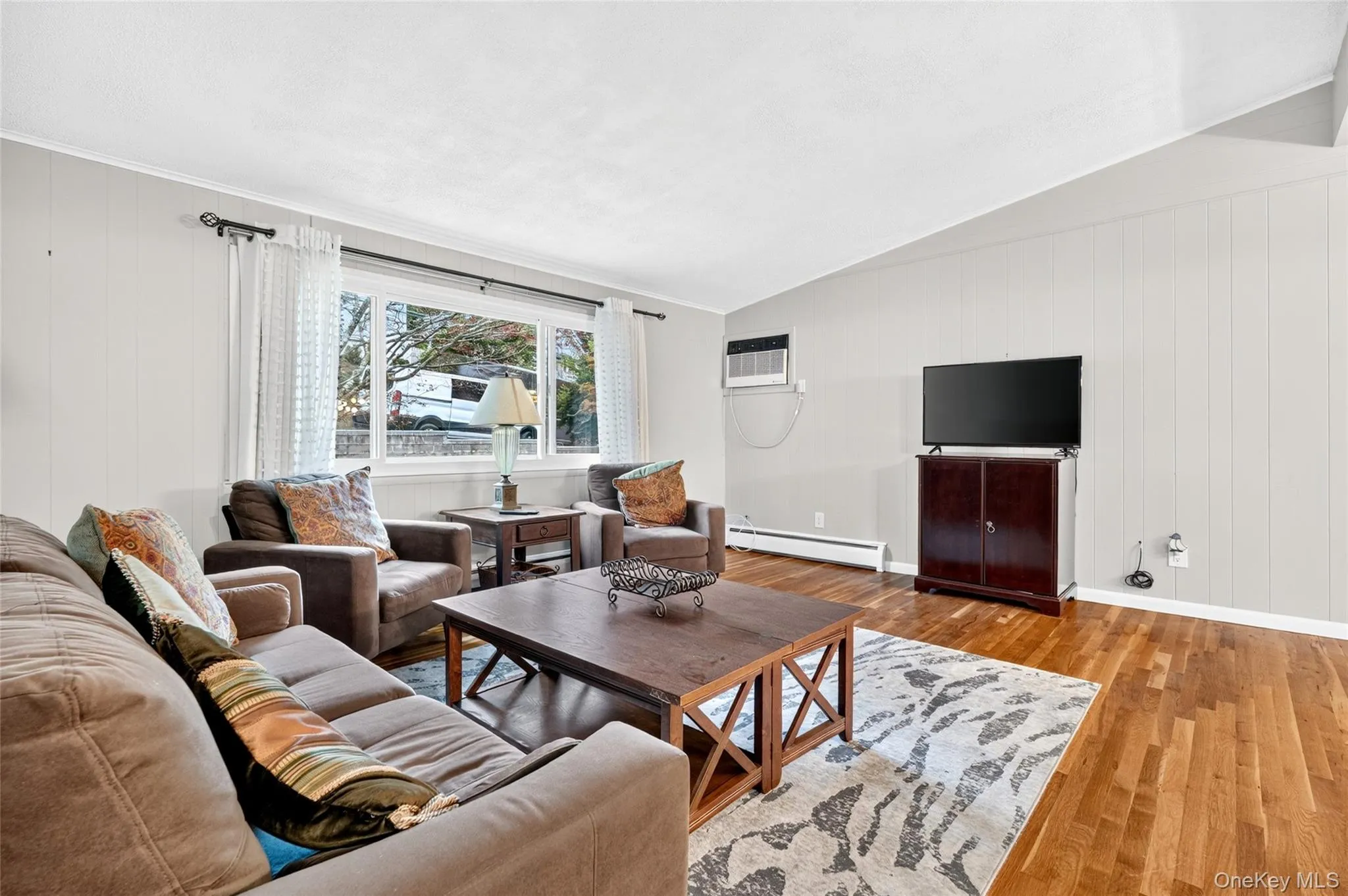 Living area featuring wood-finish flooring, white paneled walls, and a large window Living area featuring wood-finish flooring, white paneled walls, and a large window