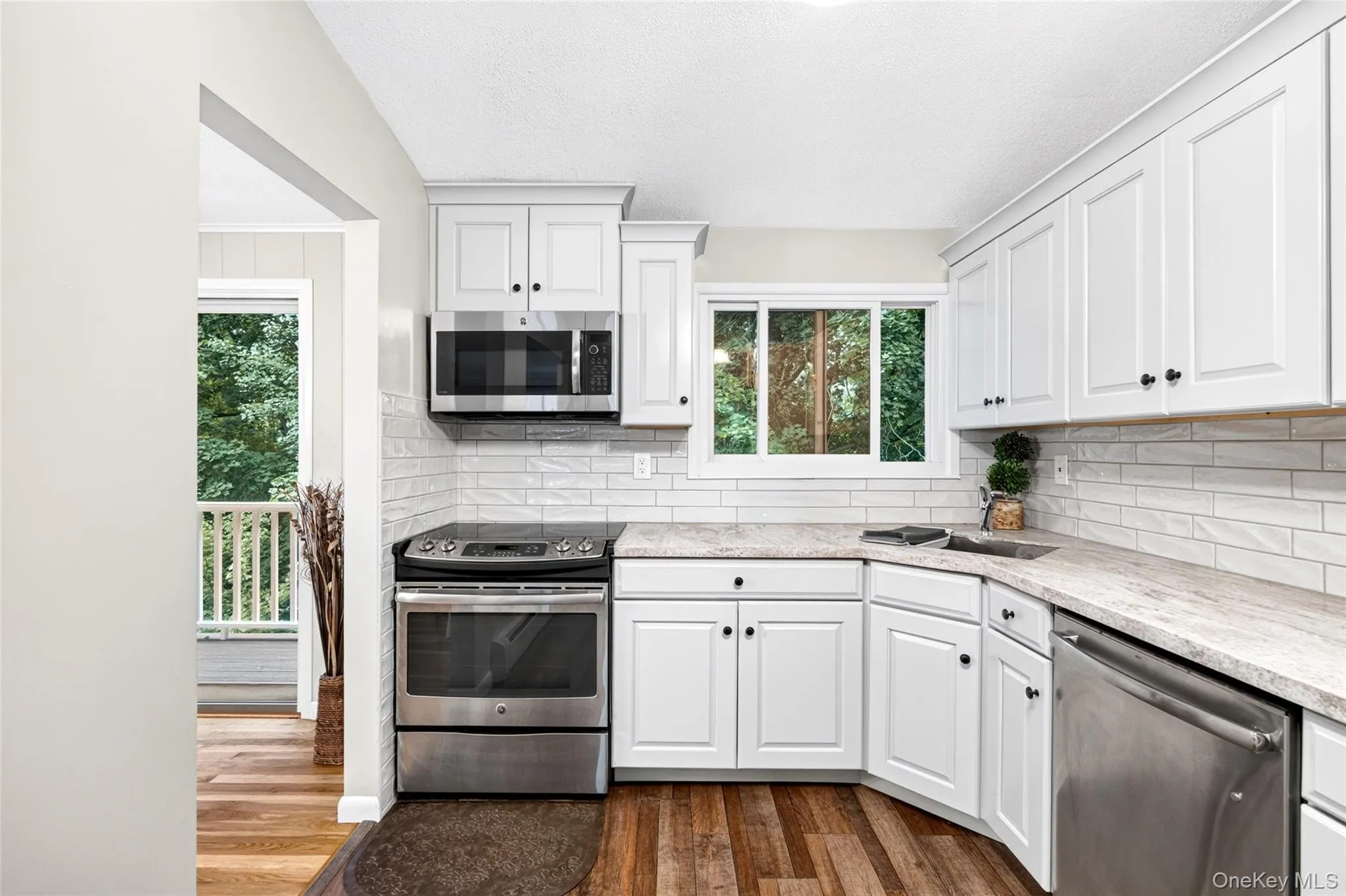 White shaker-style cabinetry with dark hardware, stainless steel appliances, wood-finish flooring, light-toned countertops, and a white subway tile backsplash White shaker-style cabinetry with dark hardware, stainless steel appliances, wood-finish flooring, light-toned countertops, and a white subway tile backsplash