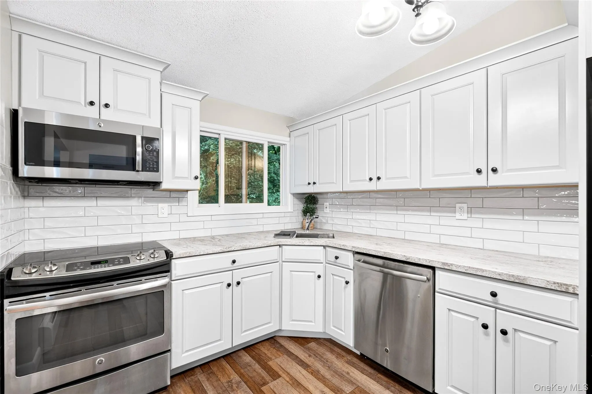 White shaker-style cabinetry, stainless steel appliances, light-toned countertops, subway tile backsplash, and wood-finish flooring White shaker-style cabinetry, stainless steel appliances, light-toned countertops, subway tile backsplash, and wood-finish flooring