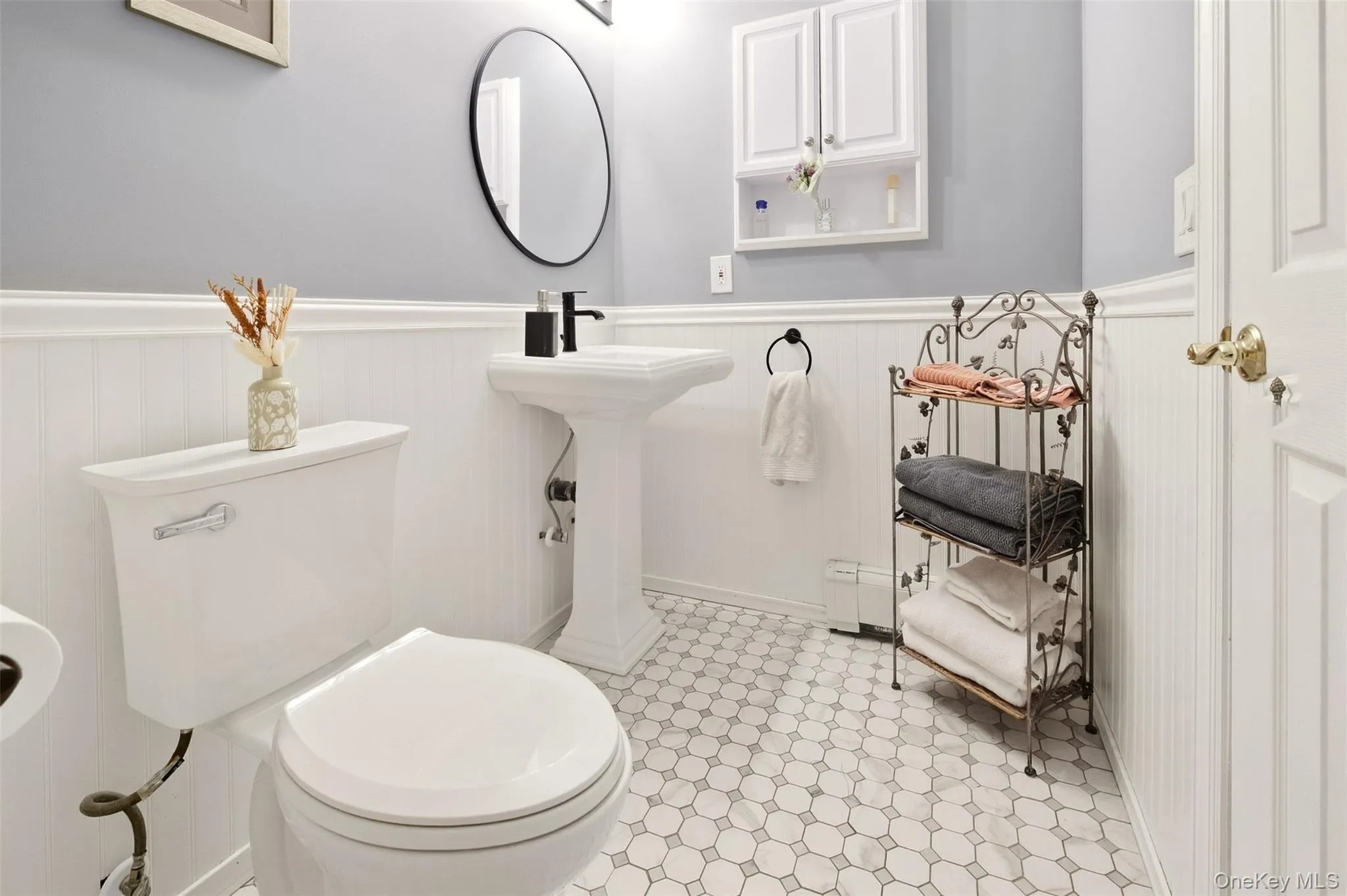 White beadboard wainscoting with a decorative trim, white hexagon tile flooring, pedestal sink with a black faucet, and an oval mirror White beadboard wainscoting with a decorative trim, white hexagon tile flooring, pedestal sink with a black faucet, and an oval mirror