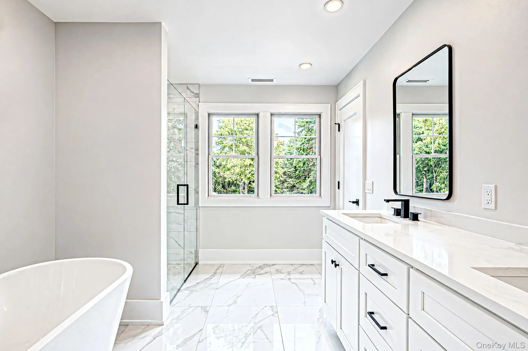 White dual vanity with integrated sinks, black fixtures, freestanding bathtub, glass-enclosed shower, and large windows White dual vanity with integrated sinks, black fixtures, freestanding bathtub, glass-enclosed shower, and large windows
