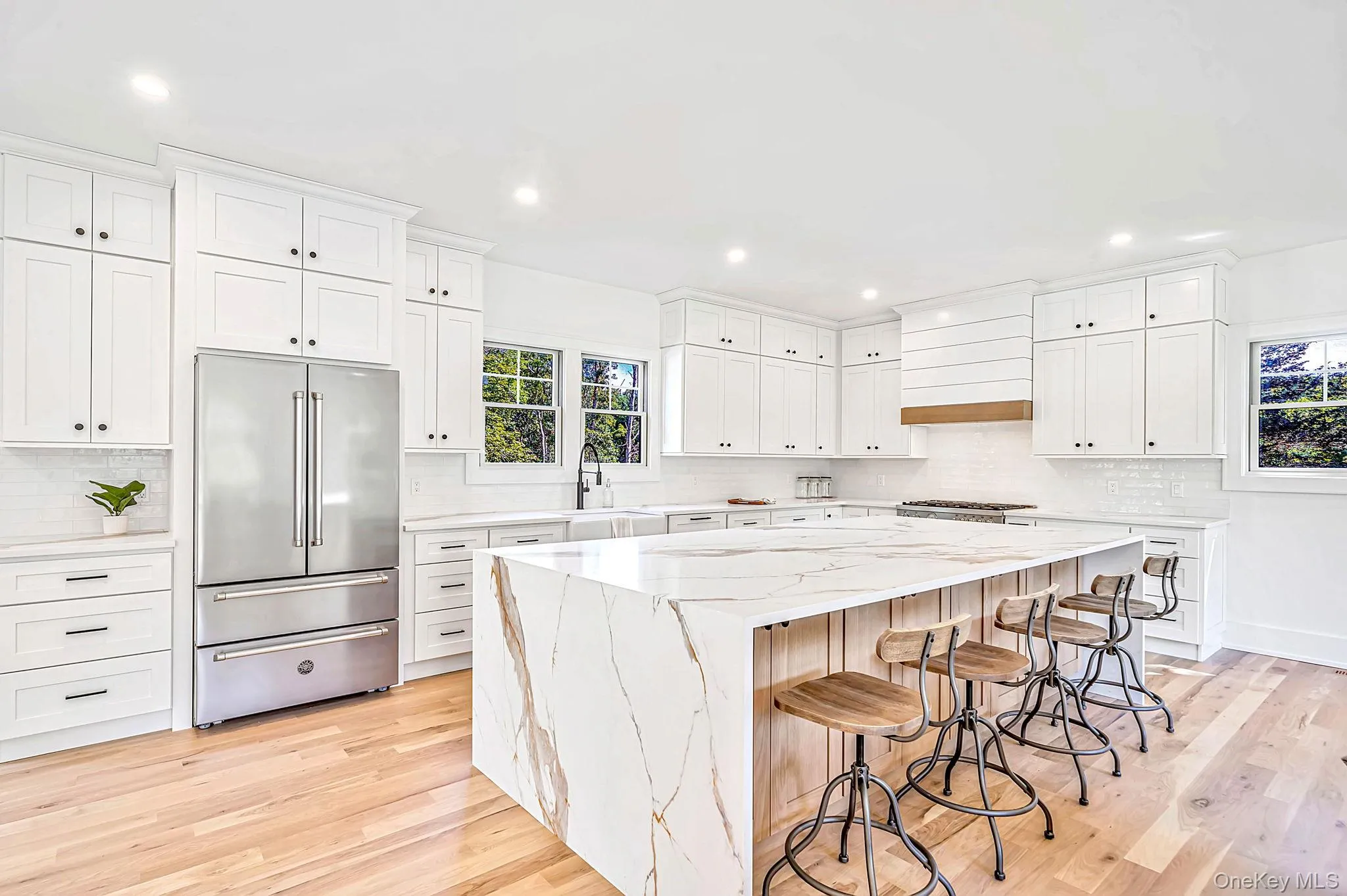 Expansive kitchen featuring a large central island with waterfall countertop, white shaker-style cabinetry, stainless steel refrigerator, and light wood-finish flooring Expansive kitchen featuring a large central island with waterfall countertop, white shaker-style cabinetry, stainless steel refrigerator, and light wood-finish flooring