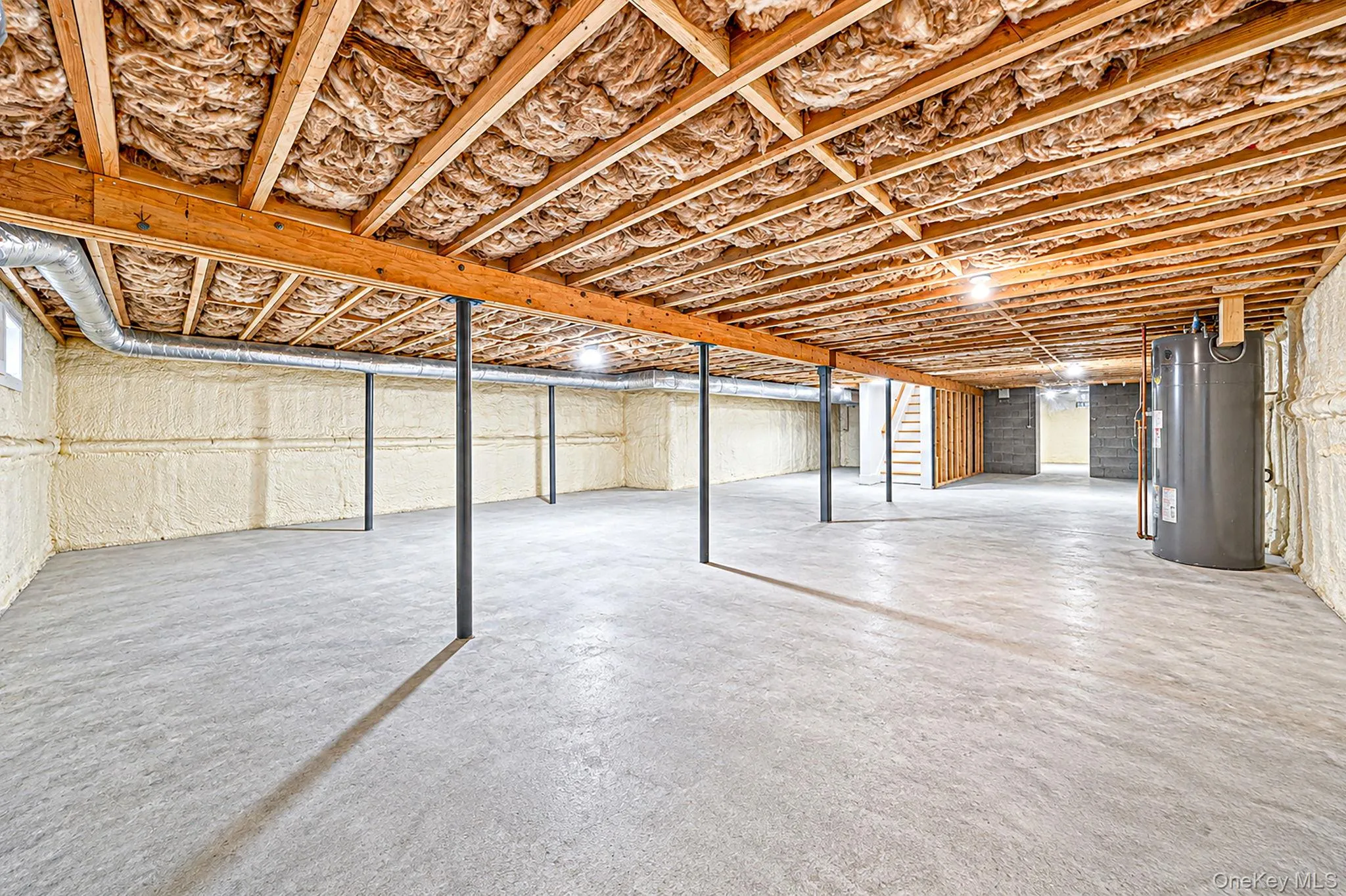 Expansive unfinished basement featuring concrete flooring, exposed wood ceiling joists with insulation, a water heater, and spray foam insulated walls Expansive unfinished basement featuring concrete flooring, exposed wood ceiling joists with insulation, a water heater, and spray foam insulated walls