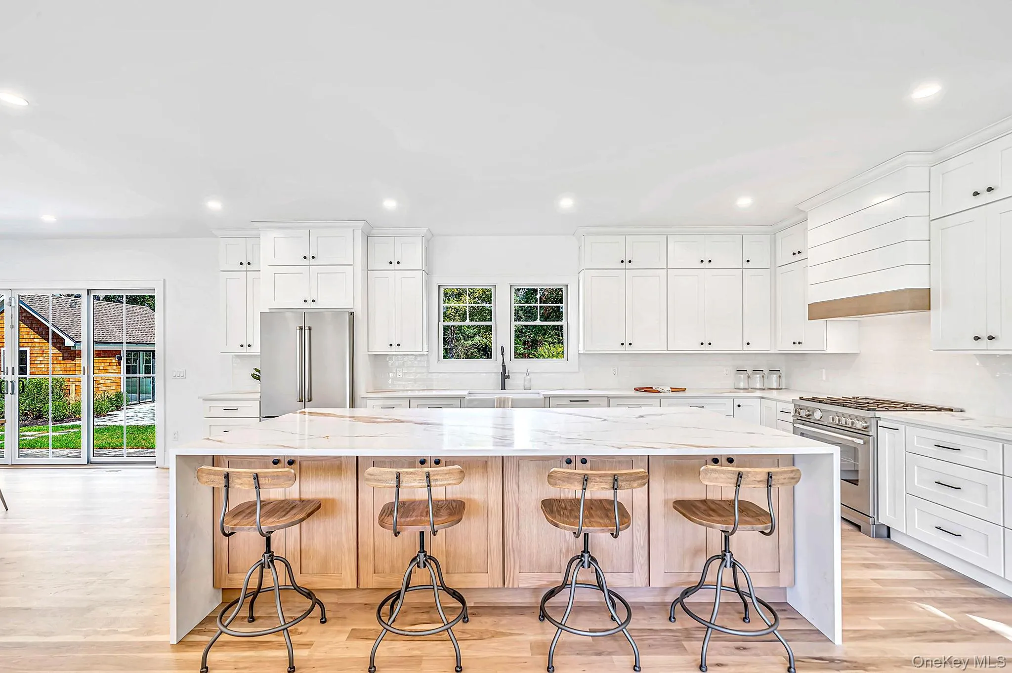Bright kitchen featuring a waterfall island, light wood-finish flooring, white shaker cabinetry, stainless steel appliances, and sliding glass doors Bright kitchen featuring a waterfall island, light wood-finish flooring, white shaker cabinetry, stainless steel appliances, and sliding glass doors