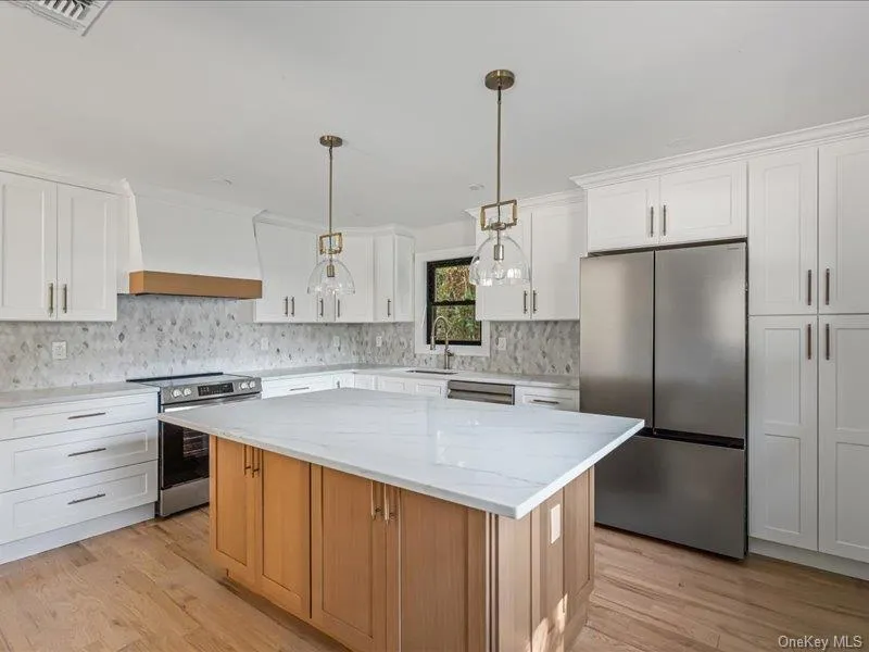 Gourmet kitchen featuring white shaker cabinetry, an expansive center island with a stone countertop, stainless steel appliances, a decorative tile backsplash, and wood-finish flooring Gourmet kitchen featuring white shaker cabinetry, an expansive center island with a stone countertop, stainless steel appliances, a decorative tile backsplash, and wood-finish flooring
