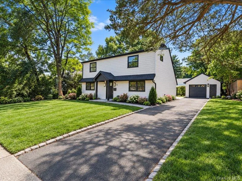 White siding exterior with black trim windows, a covered entry, an attached garage, and a paved driveway bordered by stone White siding exterior with black trim windows, a covered entry, an attached garage, and a paved driveway bordered by stone