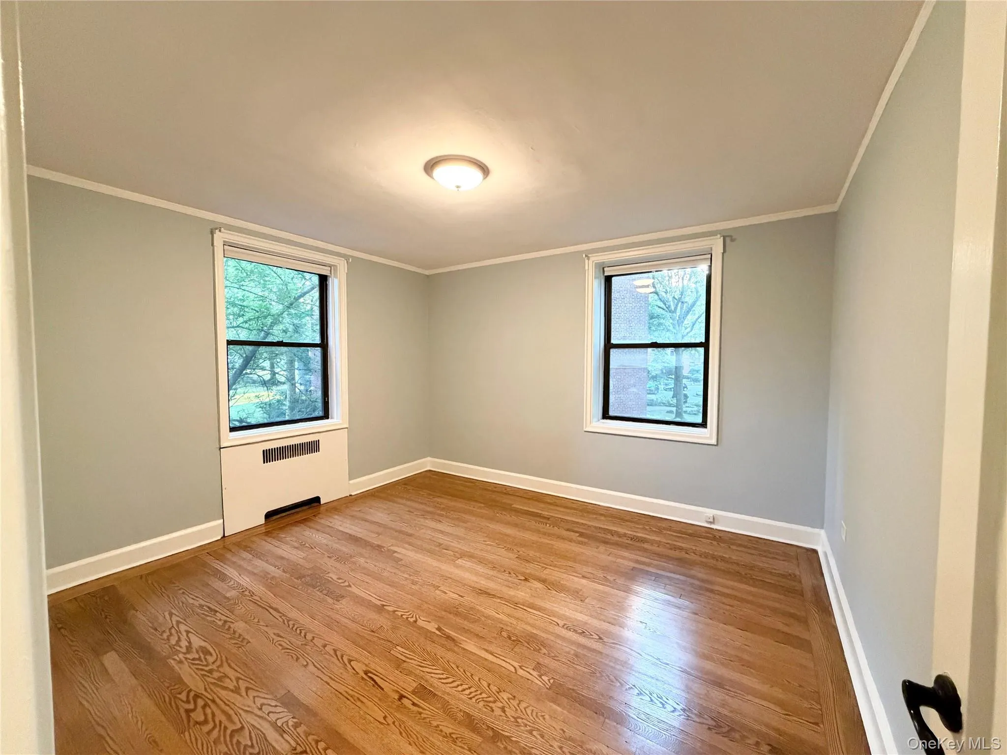 Hardwood flooring, two windows with white trim, a ceiling-mounted light fixture, and a wall-mounted heating unit. Hardwood flooring, two windows with white trim, a ceiling-mounted light fixture, and a wall-mounted heating unit.