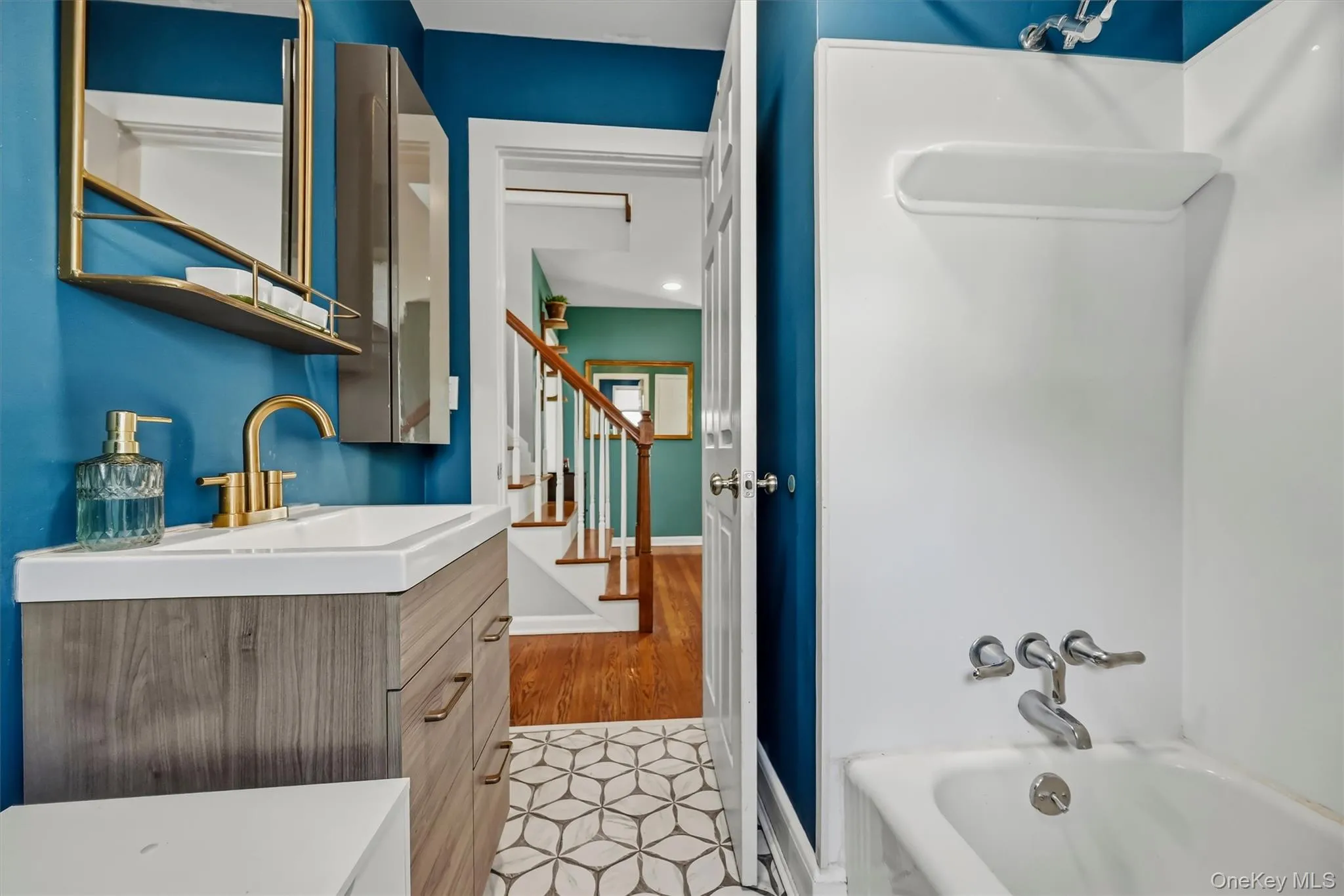Bathroom featuring a wood-finish vanity with a white countertop, gold-tone faucet, framed mirror with integrated shelving, and patterned floor tile Bathroom featuring a wood-finish vanity with a white countertop, gold-tone faucet, framed mirror with integrated shelving, and patterned floor tile