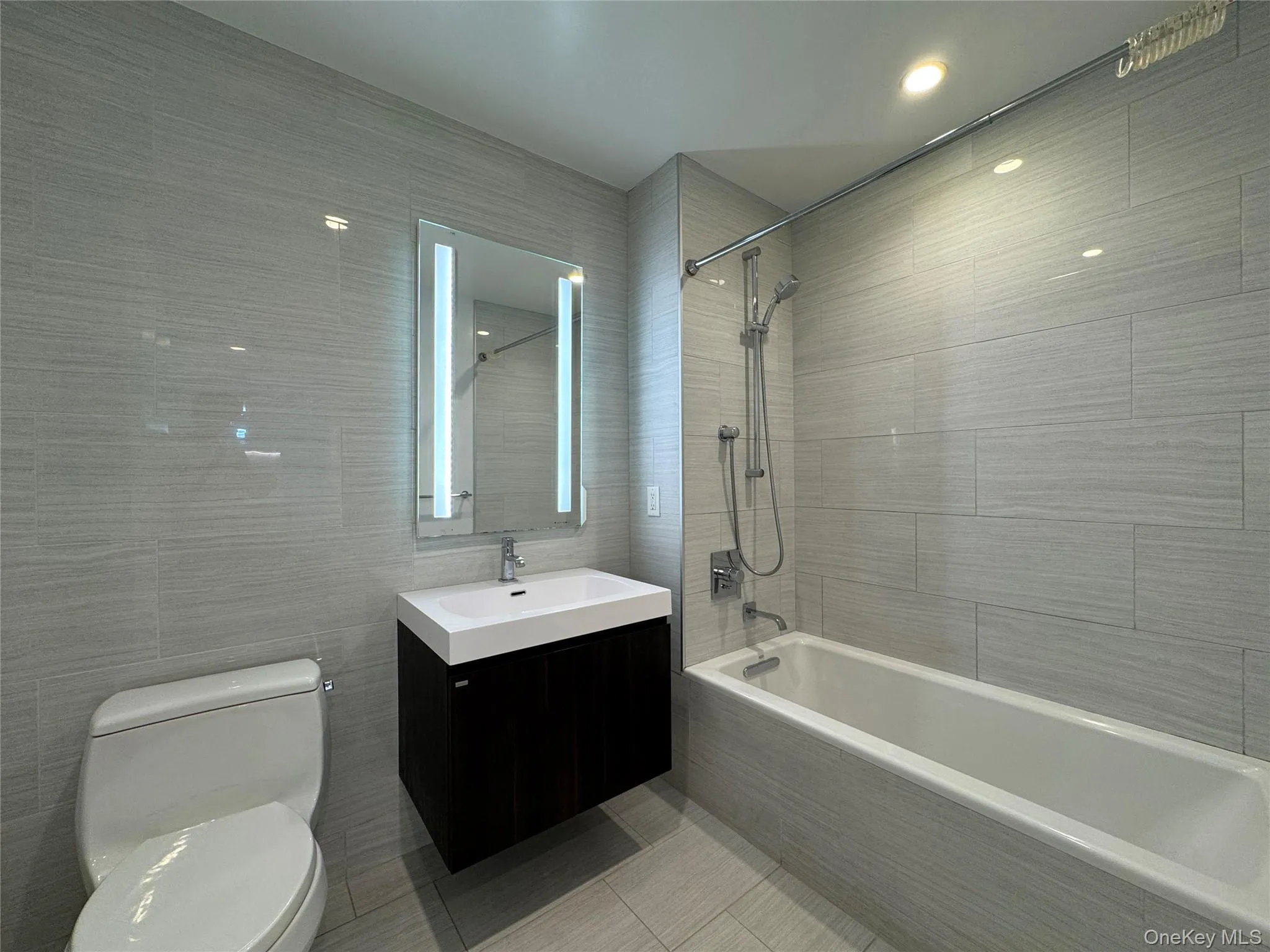 Bathroom featuring a wall-mounted vanity with integrated sink, an illuminated mirror, a bathtub with shower fixture, and light-toned tile flooring and walls Bathroom featuring a wall-mounted vanity with integrated sink, an illuminated mirror, a bathtub with shower fixture, and light-toned tile flooring and walls