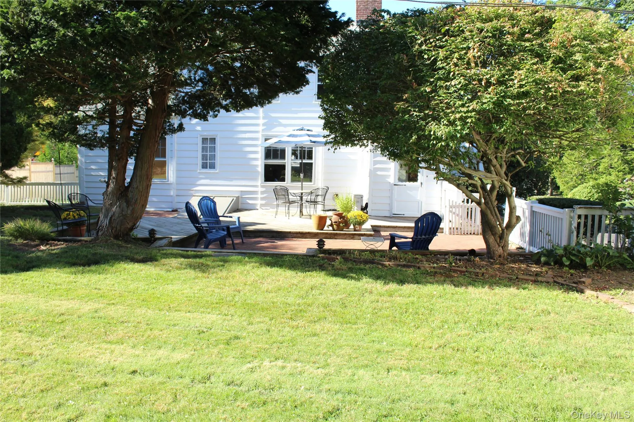Expansive lawn with mature trees, brick patio, wood decking, Bilco doors to basement and white picket fencing Expansive lawn with mature trees, brick patio, wood decking, Bilco doors to basement and white picket fencing