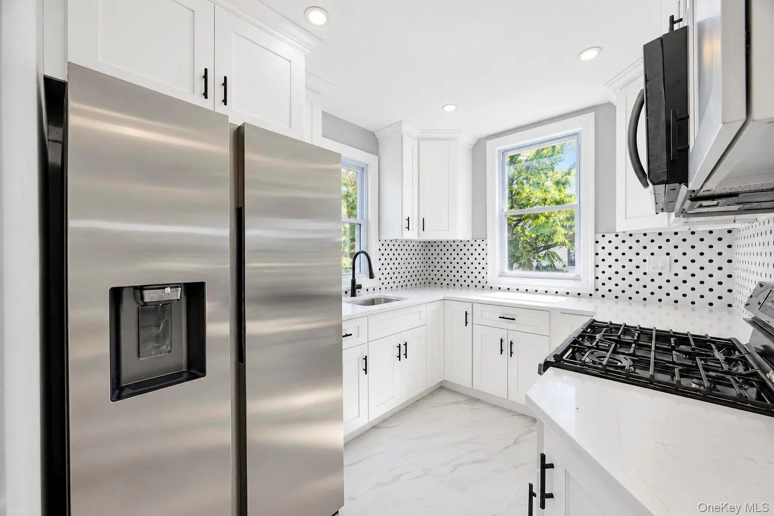 Kitchen featuring white shaker cabinetry, stainless steel appliances, black and white mosaic tile backsplash, and light-colored countertops Kitchen featuring white shaker cabinetry, stainless steel appliances, black and white mosaic tile backsplash, and light-colored countertops