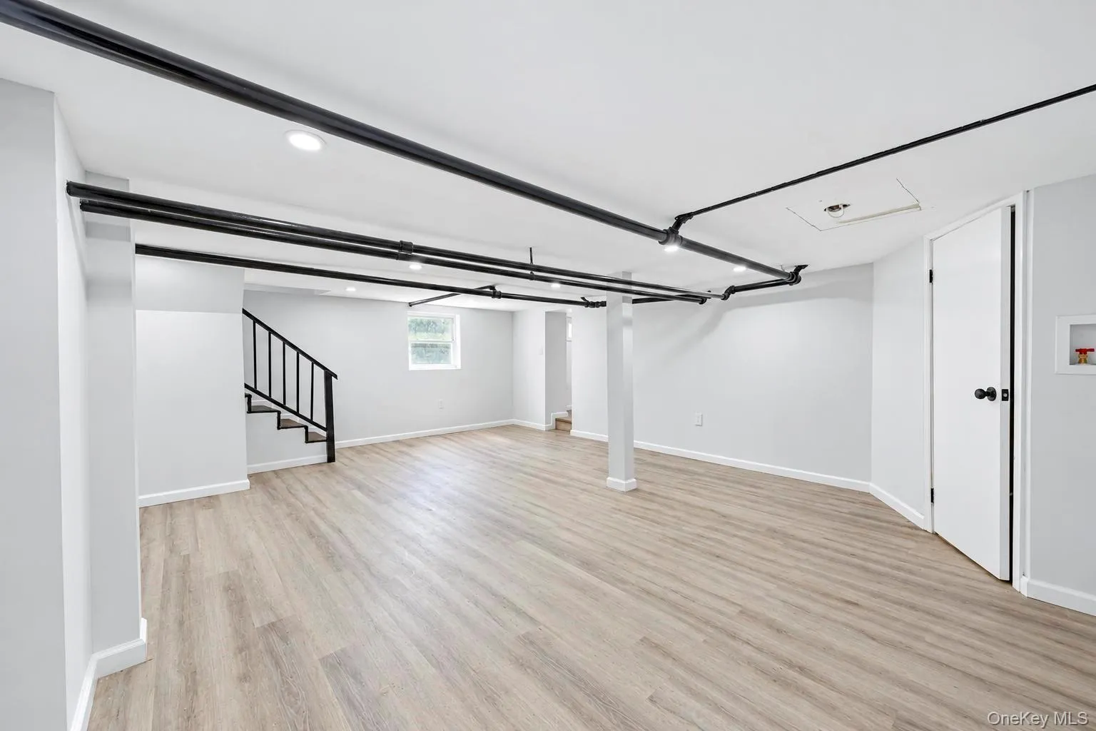 Spacious room featuring wood-finish flooring, recessed lighting, a window, and a finished staircase with metal railings Spacious room featuring wood-finish flooring, recessed lighting, a window, and a finished staircase with metal railings