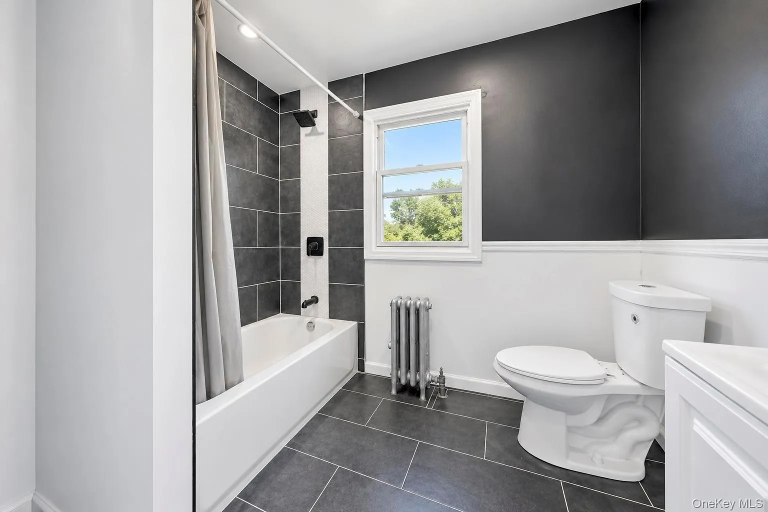 Shower and tub combination with dark tile surround, dark wood-finish flooring, white toilet, and window with exterior scenery Shower and tub combination with dark tile surround, dark wood-finish flooring, white toilet, and window with exterior scenery