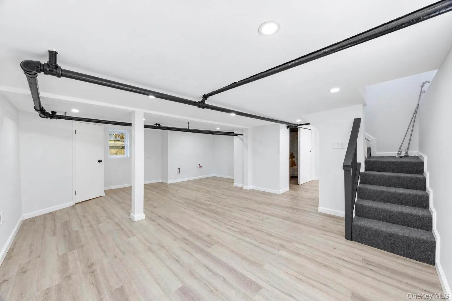 Spacious room featuring wood-finish flooring, recessed lighting, exposed ceiling pipes, and a carpeted staircase with a dark railing Spacious room featuring wood-finish flooring, recessed lighting, exposed ceiling pipes, and a carpeted staircase with a dark railing