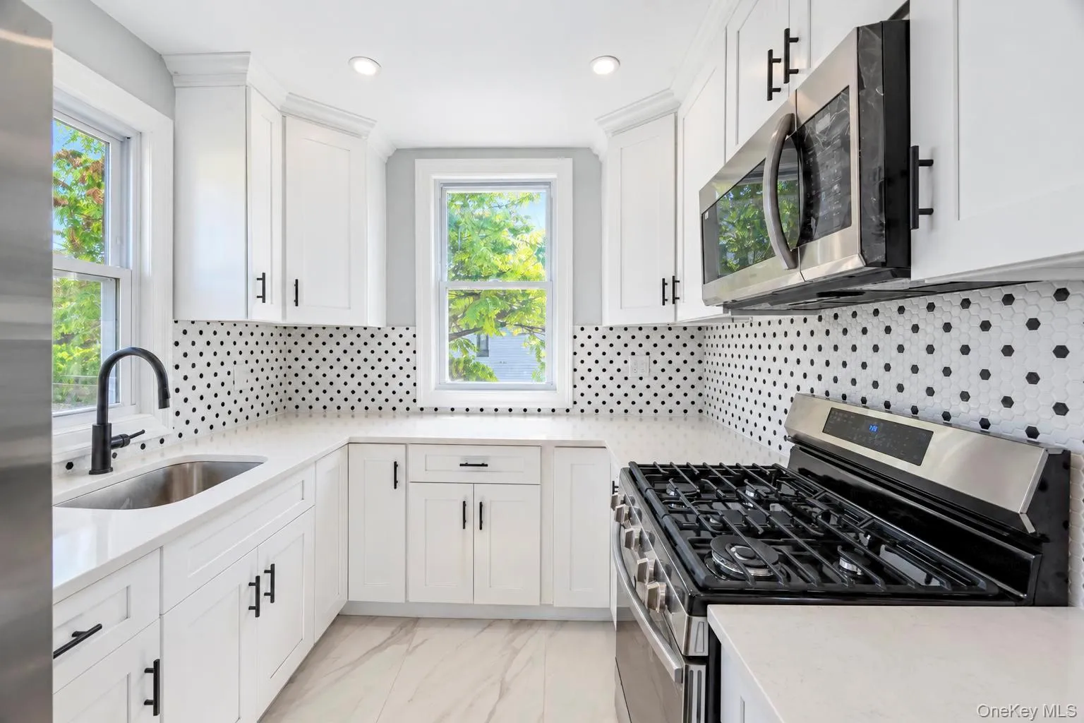 White cabinetry with black hardware, stainless steel appliances, solid surface countertops, patterned tile backsplash, and light-toned flooring White cabinetry with black hardware, stainless steel appliances, solid surface countertops, patterned tile backsplash, and light-toned flooring