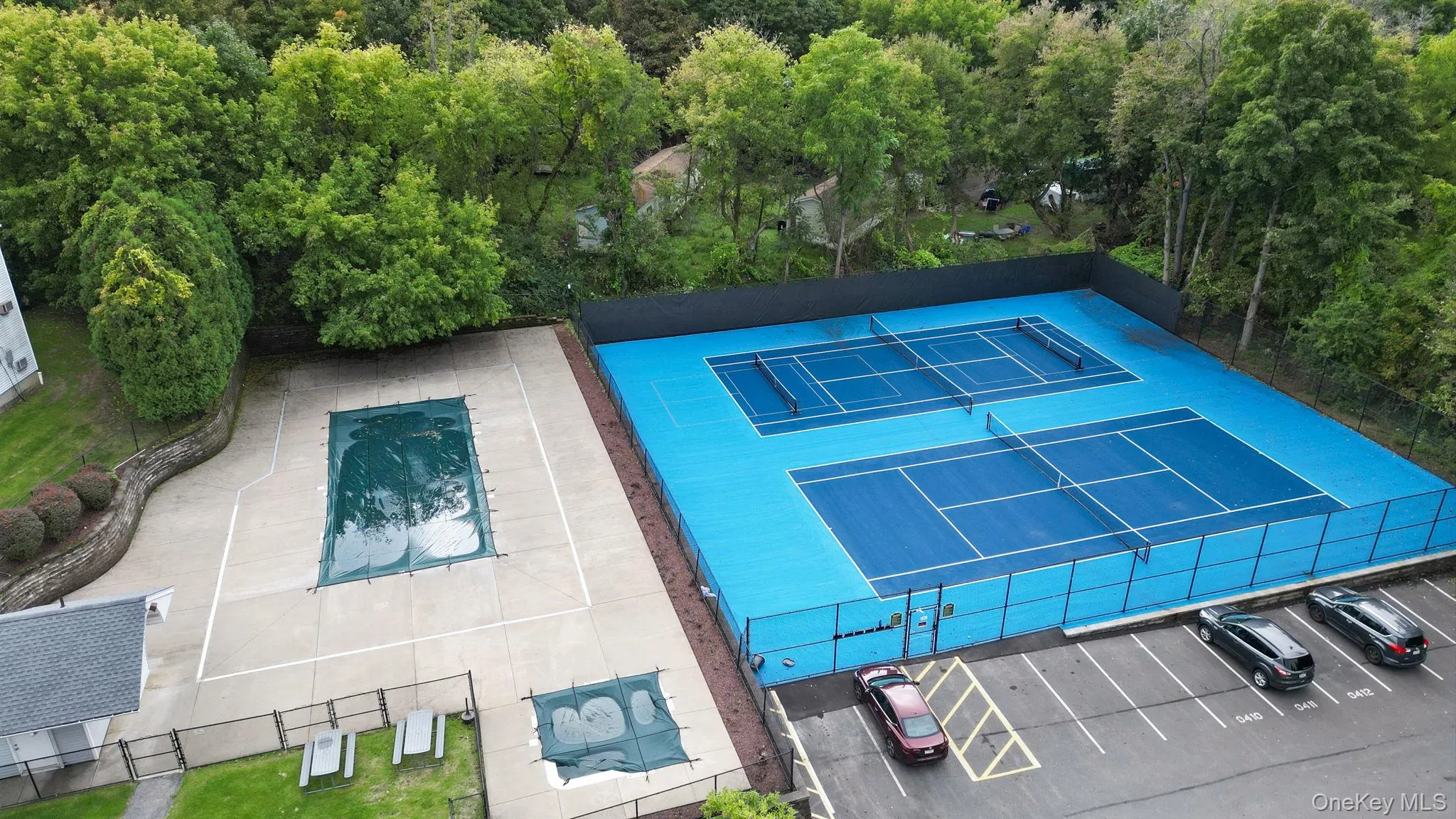 Two blue tennis courts with perimeter fencing, an adjacent covered swimming pool, a smaller covered pool, and paved parking spaces Two blue tennis courts with perimeter fencing, an adjacent covered swimming pool, a smaller covered pool, and paved parking spaces