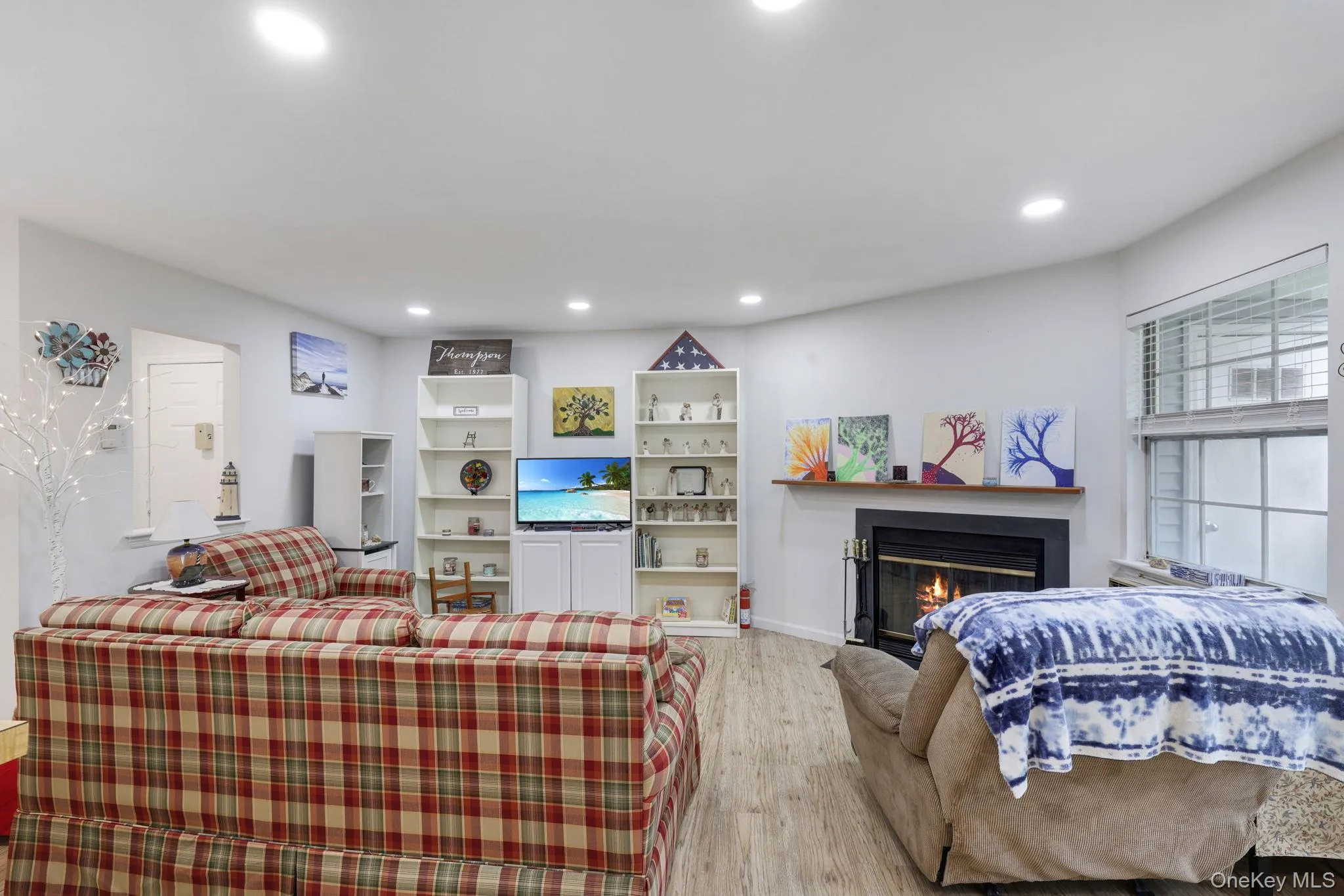 Recessed lighting illumination, wood-finish flooring, built-in shelving, corner fireplace with wood mantel, and large window with horizontal blinds Recessed lighting illumination, wood-finish flooring, built-in shelving, corner fireplace with wood mantel, and large window with horizontal blinds