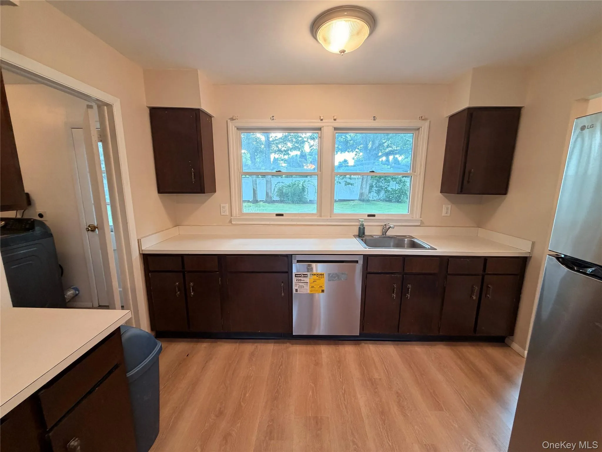 Wood-finish flooring, dark wood cabinetry, light countertops, stainless steel sink, and a large window Wood-finish flooring, dark wood cabinetry, light countertops, stainless steel sink, and a large window