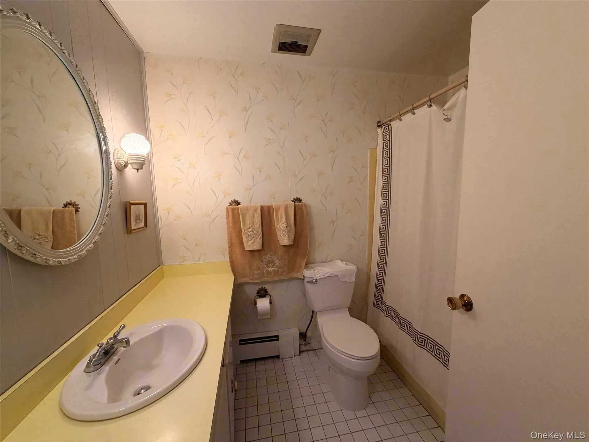 Oval vanity mirror with ornate frame, wall-mounted light fixture, integrated sink, and white ceramic tile flooring Oval vanity mirror with ornate frame, wall-mounted light fixture, integrated sink, and white ceramic tile flooring