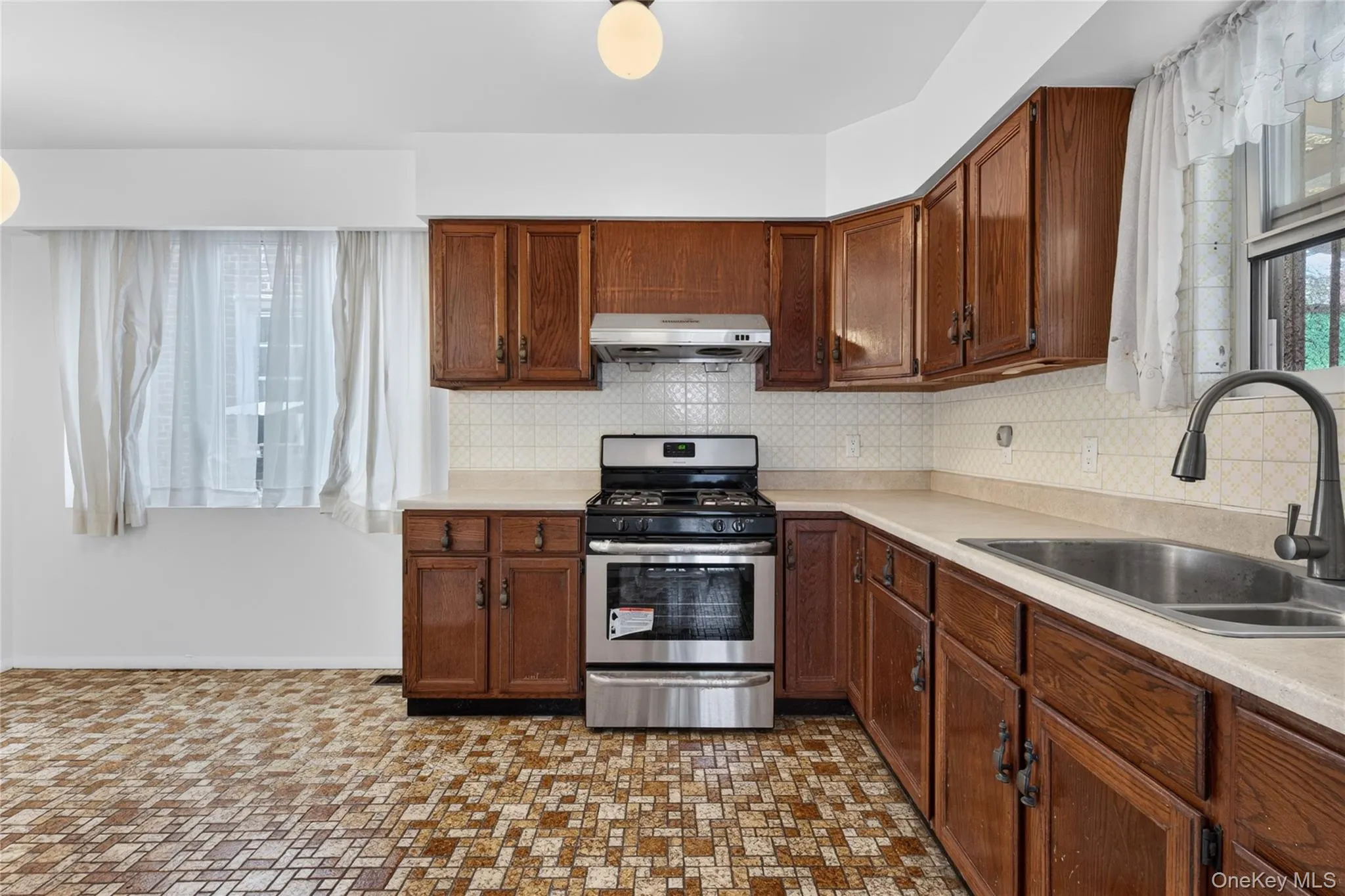 Kitchen featuring wood cabinetry, stainless steel range, a coordinating range hood, a tile backsplash, and a stainless steel sink with a dark-finish faucet Kitchen featuring wood cabinetry, stainless steel range, a coordinating range hood, a tile backsplash, and a stainless steel sink with a dark-finish faucet