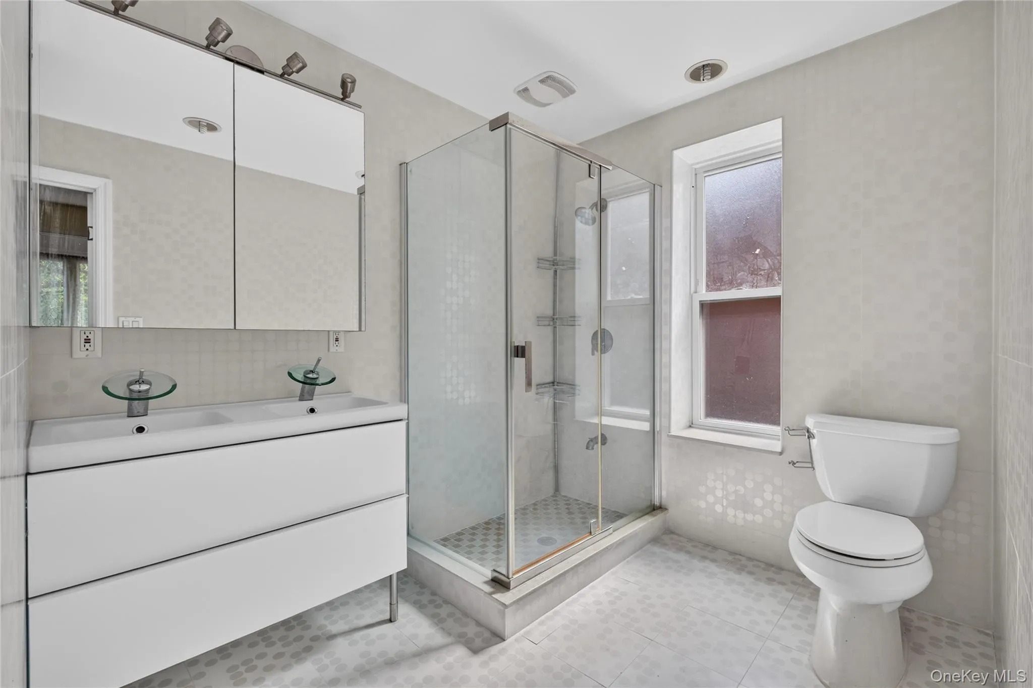 Dual basin vanity with integrated sinks, glass-enclosed shower, white tile flooring, and a single window Dual basin vanity with integrated sinks, glass-enclosed shower, white tile flooring, and a single window
