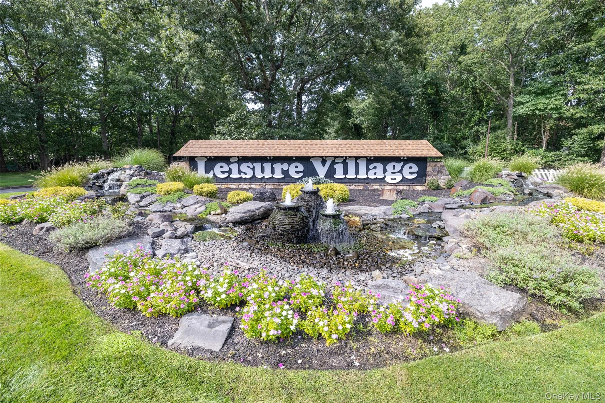 Stone-faced entrance sign with a shingled roof, surrounded by diverse plantings, a multi-tiered rock waterfall feature, and a manicured lawn Stone-faced entrance sign with a shingled roof, surrounded by diverse plantings, a multi-tiered rock waterfall feature, and a manicured lawn