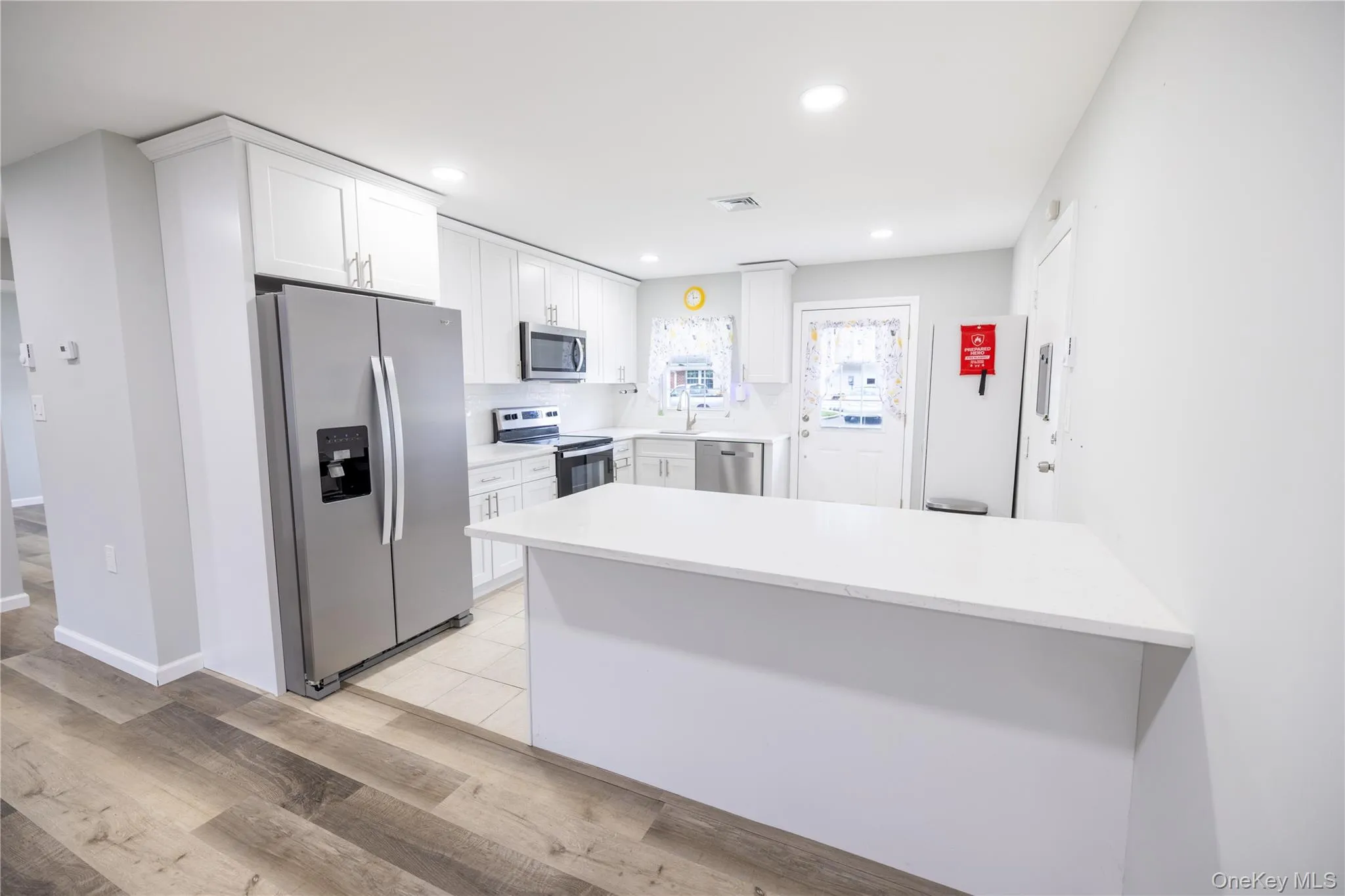 Modern kitchen featuring white shaker cabinetry, stainless steel appliances, a center island with a white countertop, and wood-finish flooring Modern kitchen featuring white shaker cabinetry, stainless steel appliances, a center island with a white countertop, and wood-finish flooring