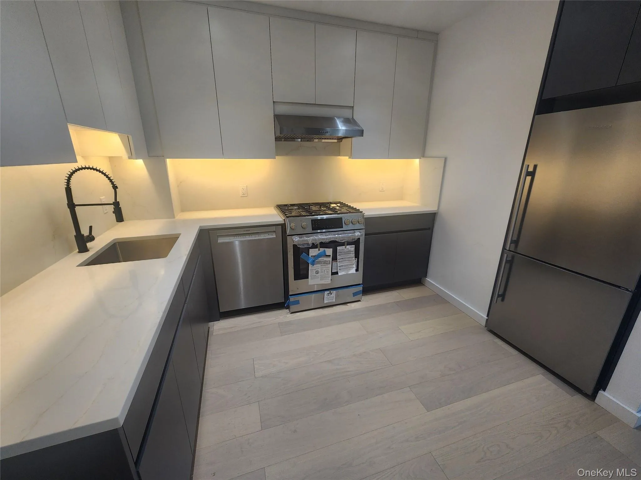 Modern kitchen featuring stainless steel appliances, light-toned wood-finish flooring, white countertops, a stainless steel sink with a matte black faucet, and two-tone cabinetry with under-cabinet lighting Modern kitchen featuring stainless steel appliances, light-toned wood-finish flooring, white countertops, a stainless steel sink with a matte black faucet, and two-tone cabinetry with under-cabinet lighting