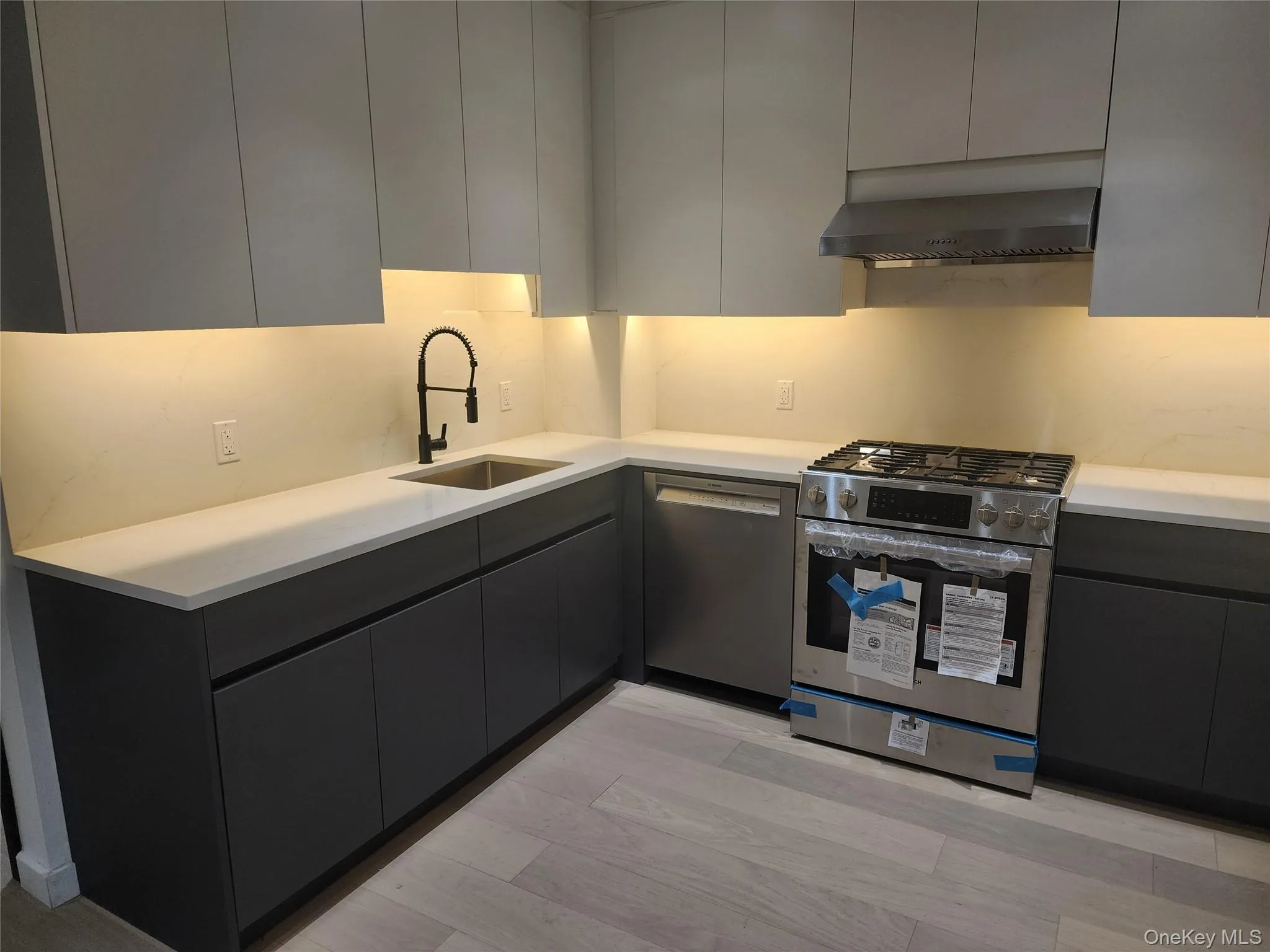 Modern kitchen featuring flat-panel cabinetry, light-toned countertops, under-cabinet lighting, stainless steel appliances, and wood-finish flooring Modern kitchen featuring flat-panel cabinetry, light-toned countertops, under-cabinet lighting, stainless steel appliances, and wood-finish flooring