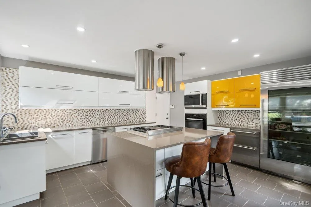 Sleek kitchen featuring a center island with integrated cooktop, mosaic tile backsplash, white and yellow high-gloss cabinetry, stainless steel appliances, and recessed lighting Sleek kitchen featuring a center island with integrated cooktop, mosaic tile backsplash, white and yellow high-gloss cabinetry, stainless steel appliances, and recessed lighting